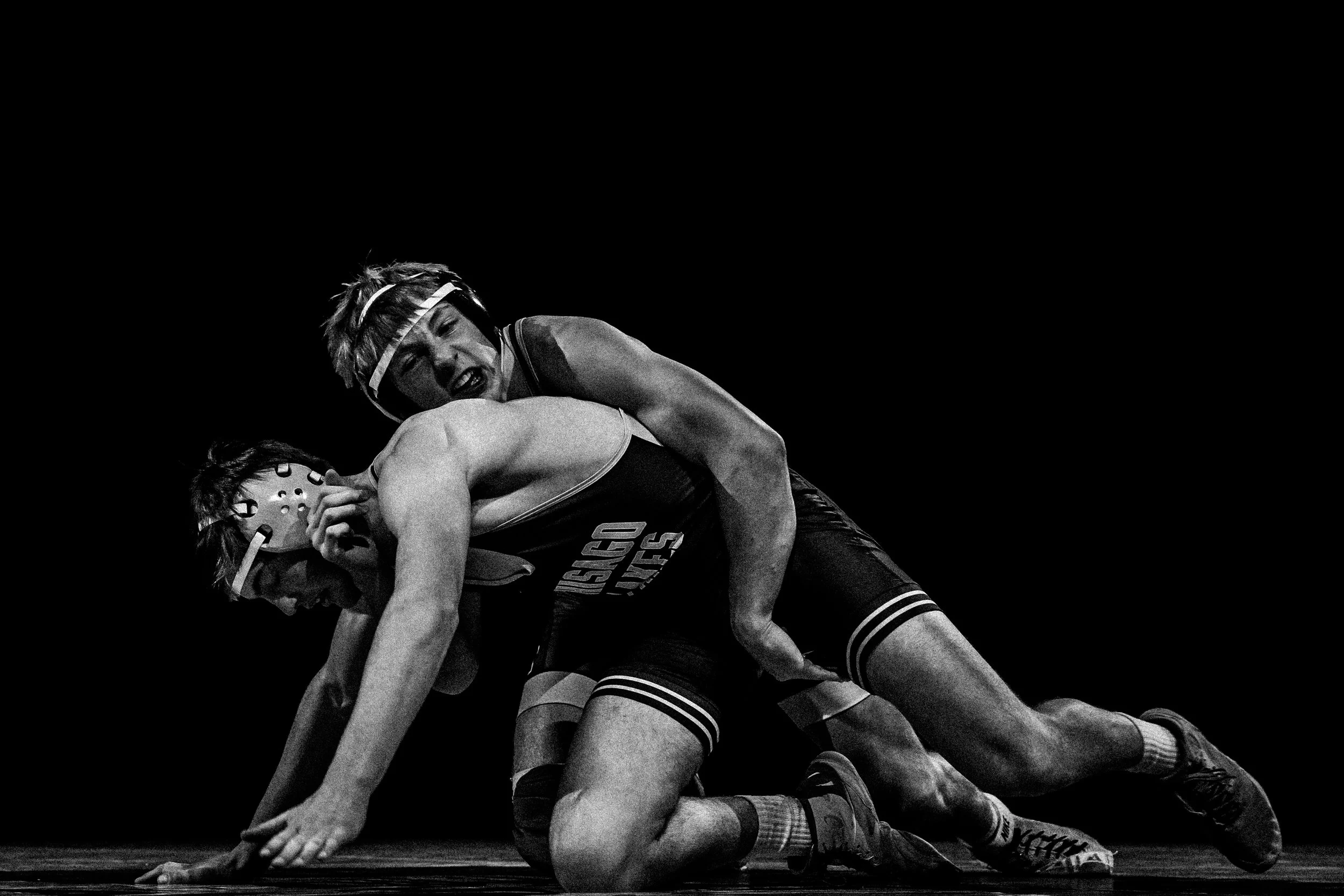Black and white photo of two women engaged in a wrestling match, one on top of the other, both in athletic attire, with a dark background.