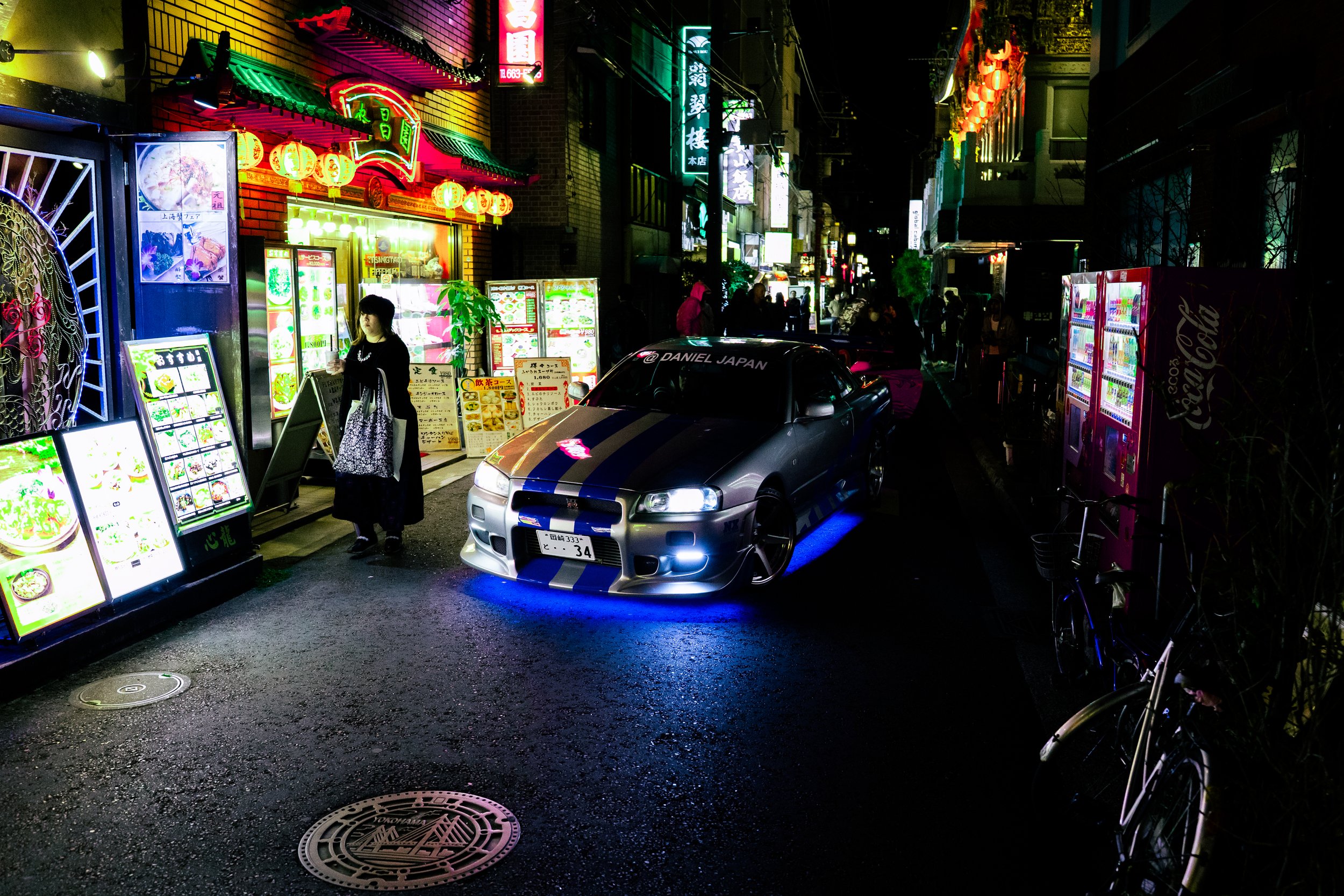 Night scene on a brightly lit city street with colorful neon signs in Chinese characters. A woman stands near menu boards outside a restaurant, and a silver sports car is parked on the street. Bicycles lean against the sidewalk, and people are walkin