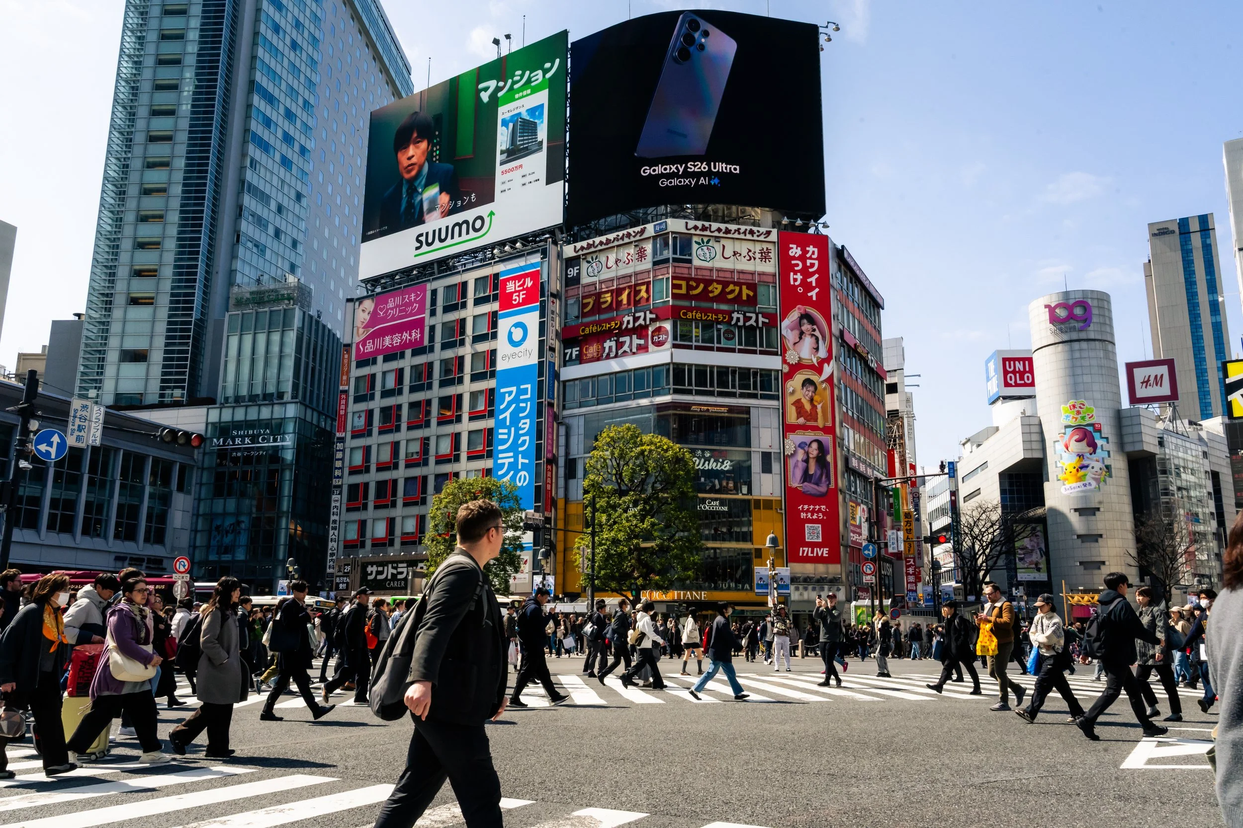A busy city crosswalk in Tokyo, Japan, with numerous pedestrians walking on the crosswalk and tall buildings in the background displaying large electronic advertisements.