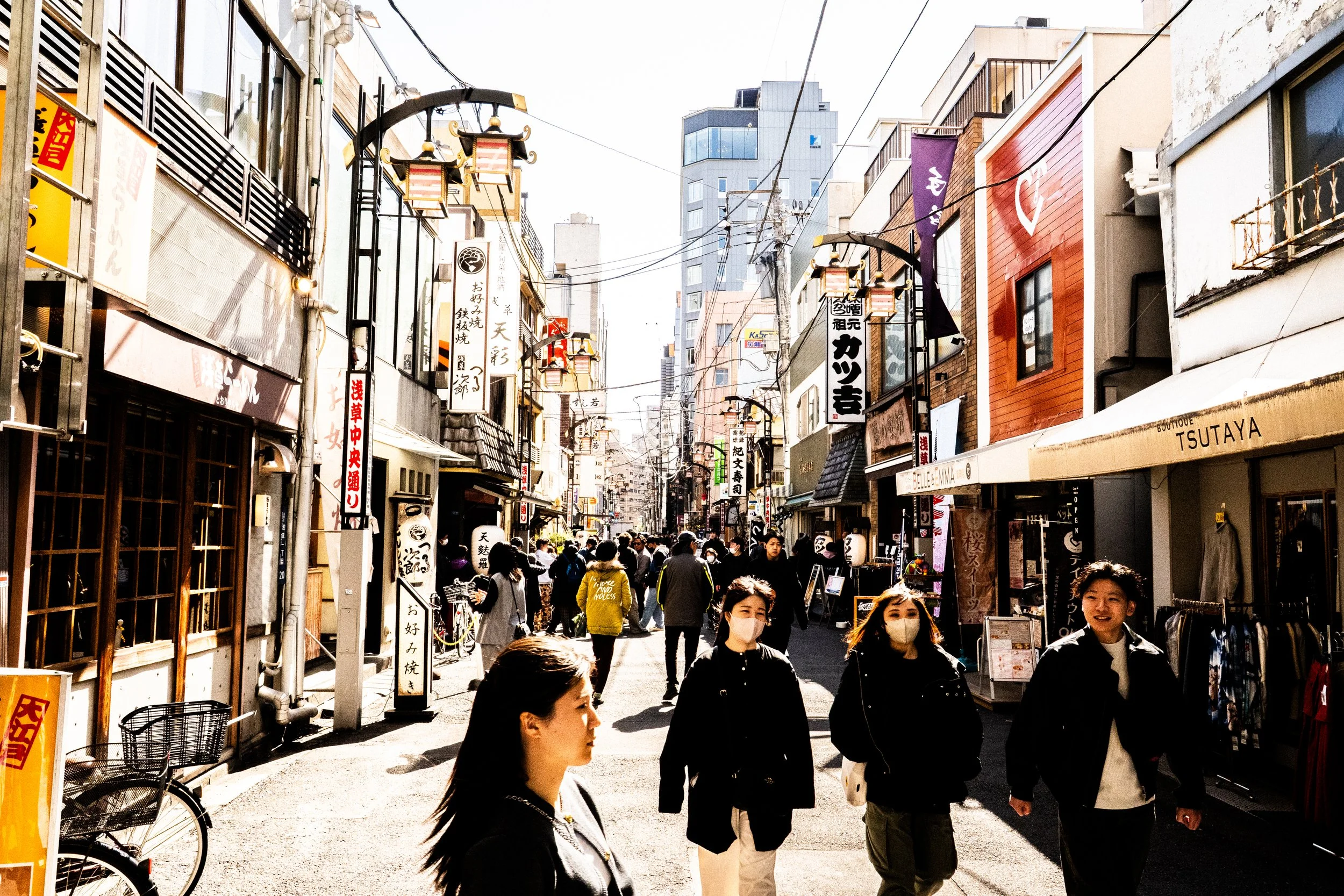 Street scene in Japan with people walking, shop signs in Japanese, and tall buildings in the background.