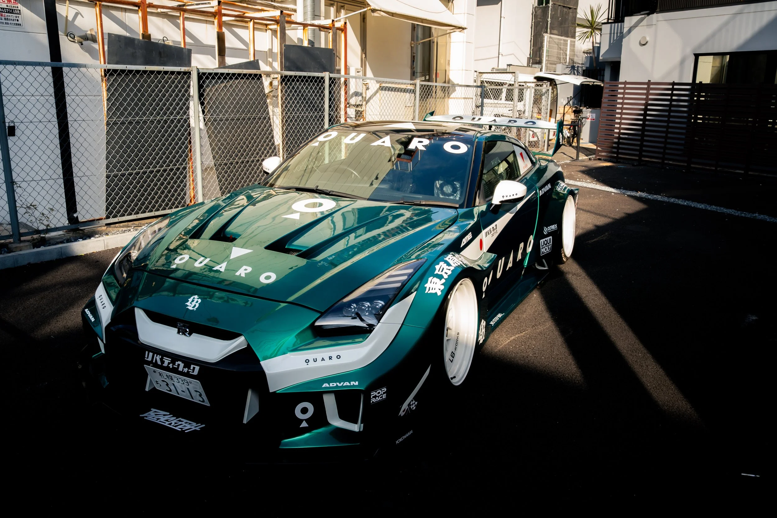 A dark green and white racing car with various sponsor logos and decals, parked on a city street next to a chain-link fence and buildings in the background.