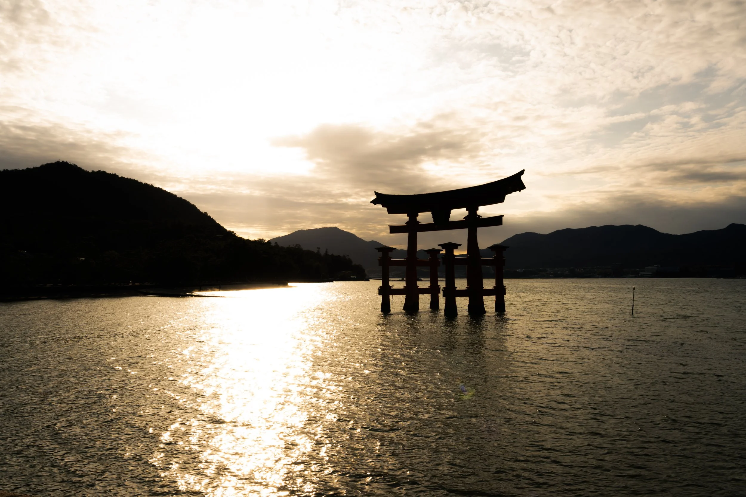 Silhouette of a traditional Japanese torii gate standing in water during sunset, with mountains and cloudy sky in the background.
