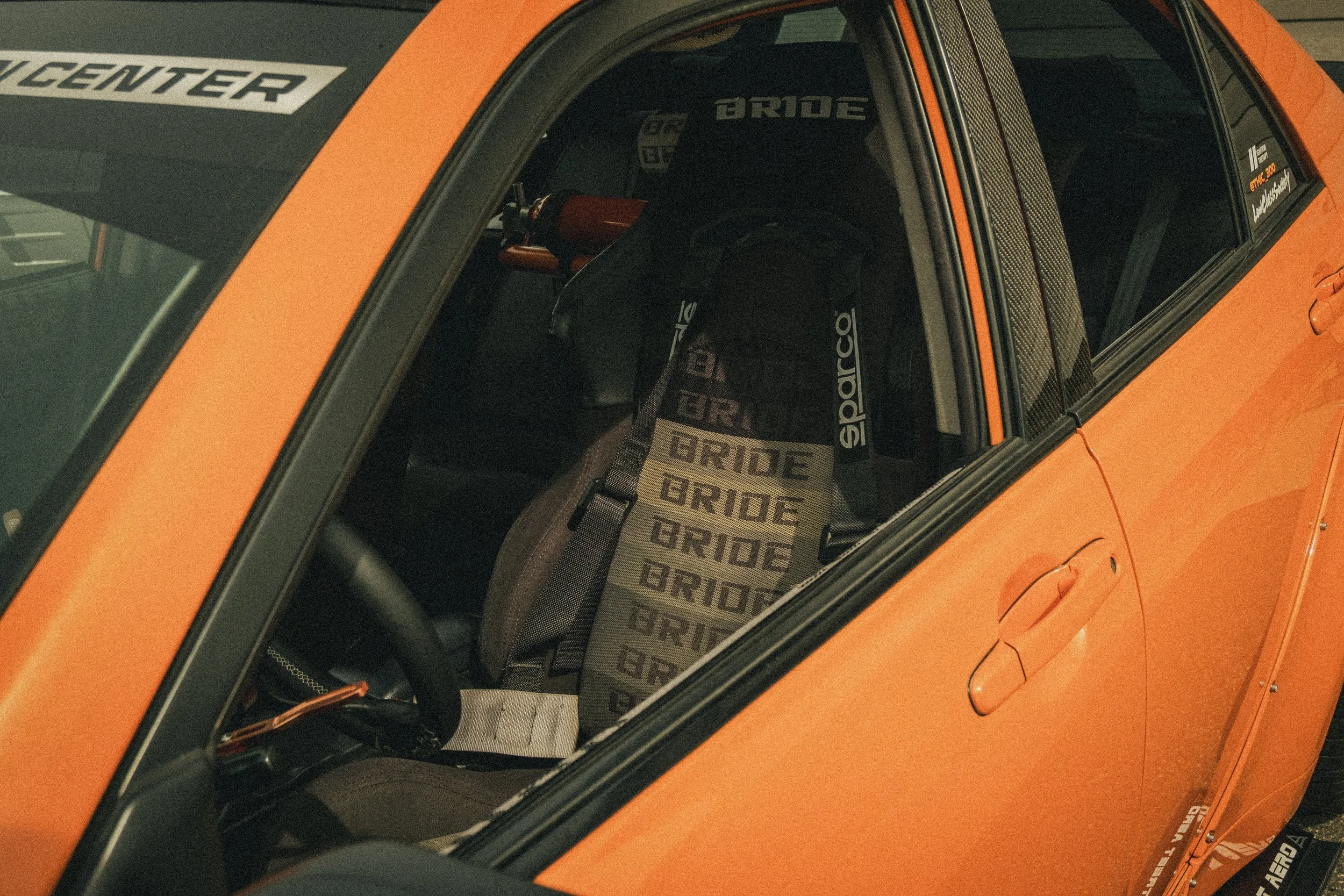 A sports car with an orange body and black roof, featuring a racing seat with 'BRIDE' written on the headrest, and the interior visible through the front window.