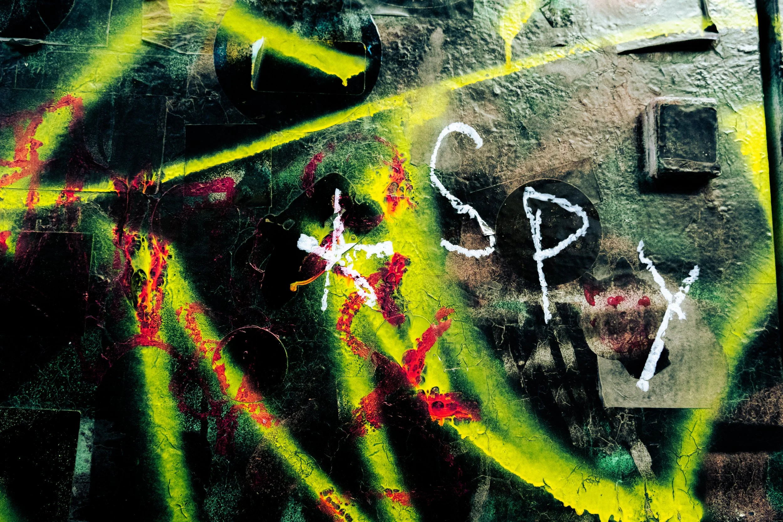 Close-up of colorful abstract street art or graffiti on a wall, featuring vibrant yellow, red, black, and green splashes and streaks with a textured surface.