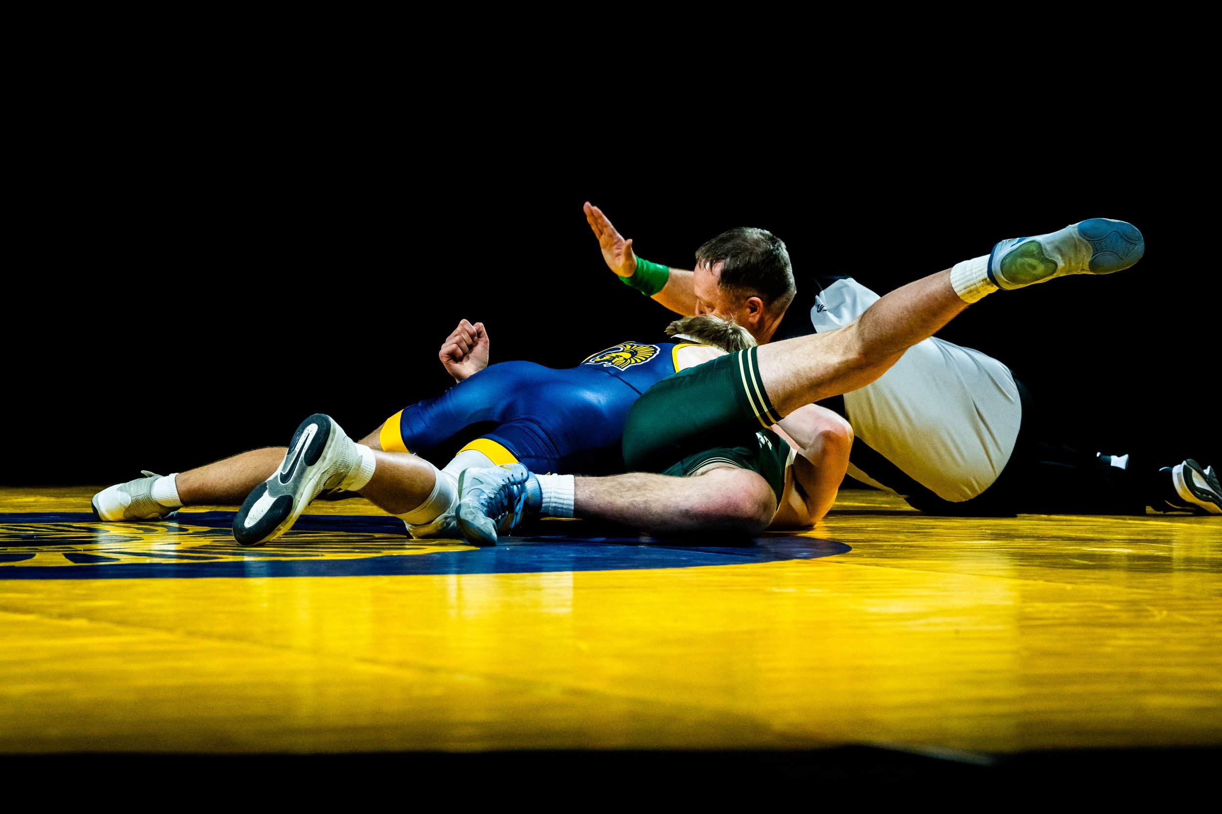 Two wrestlers on a yellow wrestling mat in a match, one in black and white and the other in blue and yellow, with a referee nearby, all against a black background.