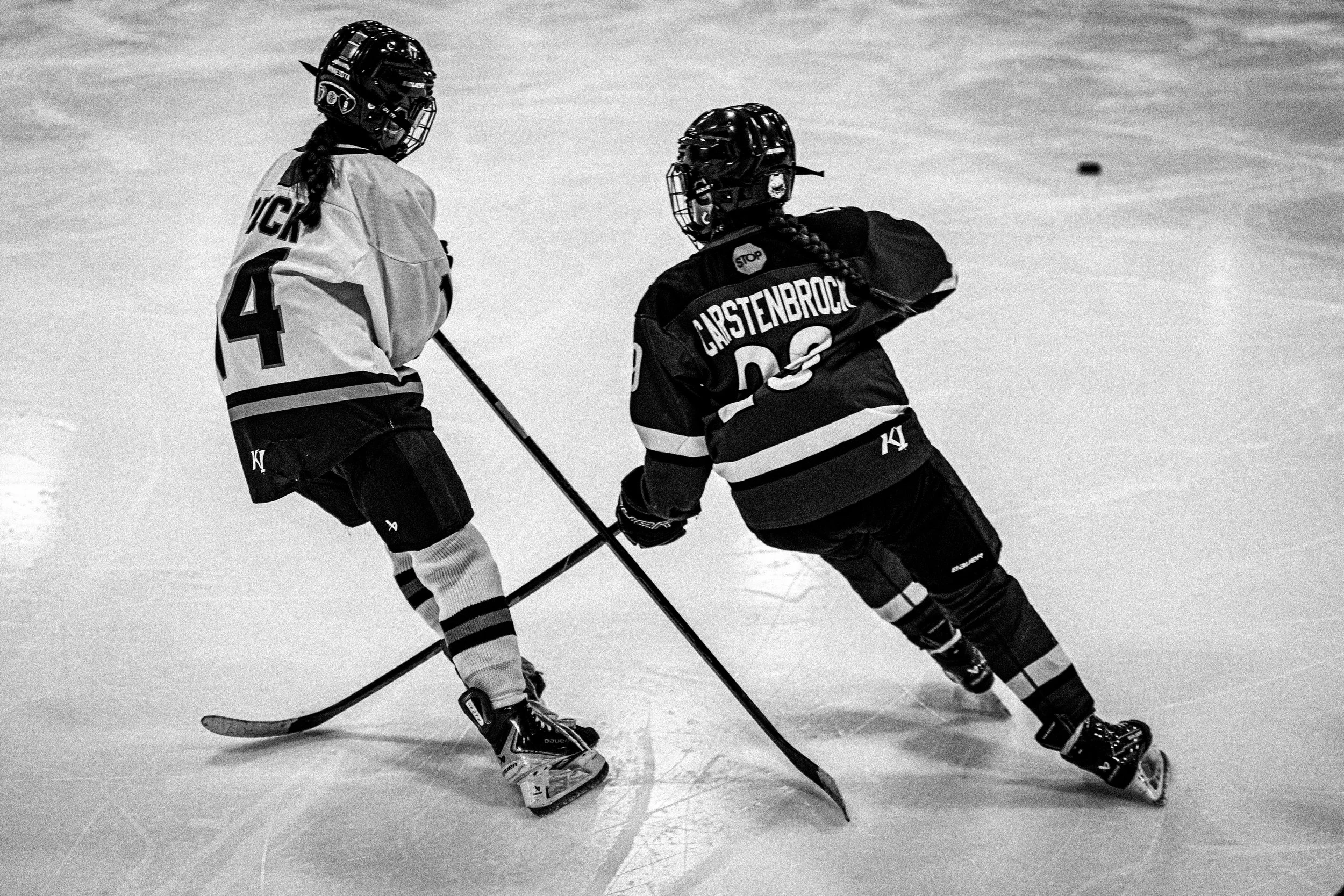 Two ice hockey players in black and white uniforms face each other on the ice rink, wearing helmets, gloves, and skates, with their sticks on the ice.