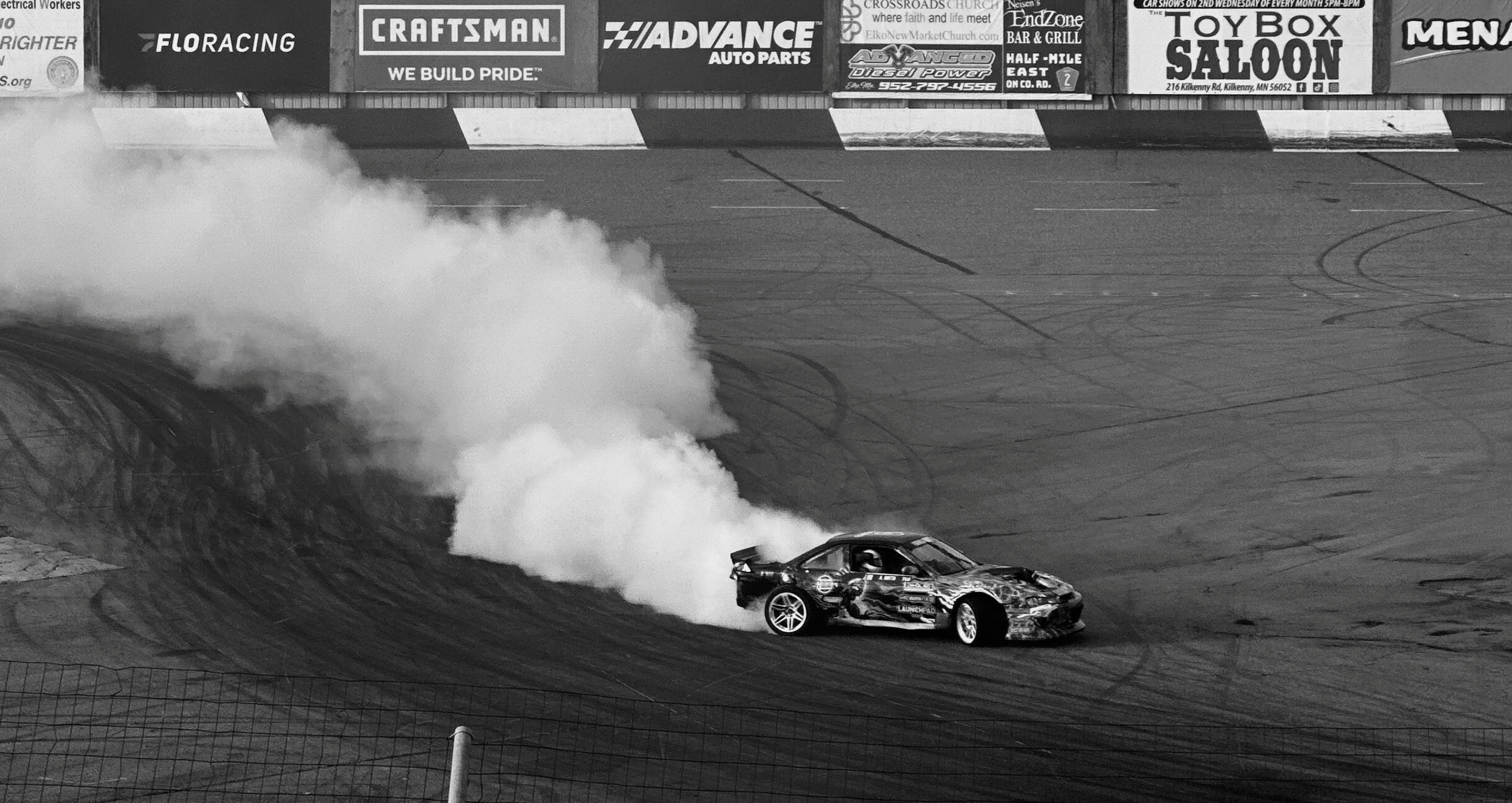 A race car drifting on a racetrack with tire smoke. The background has billboards and signs, including brands like Craftsman and Advance Auto Parts.
