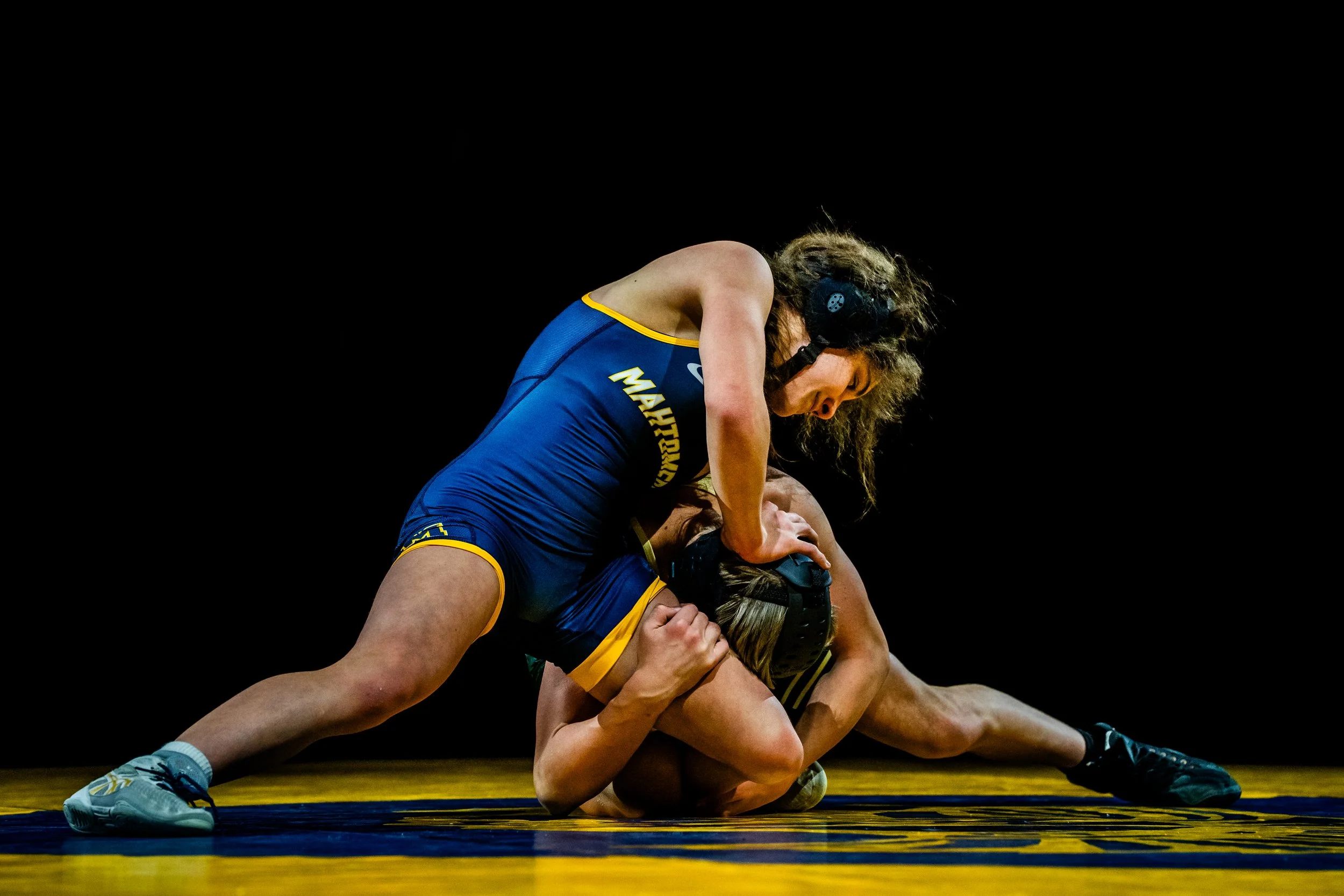 Two female wrestlers in blue uniforms competing on a wrestling mat, engaged in a match.