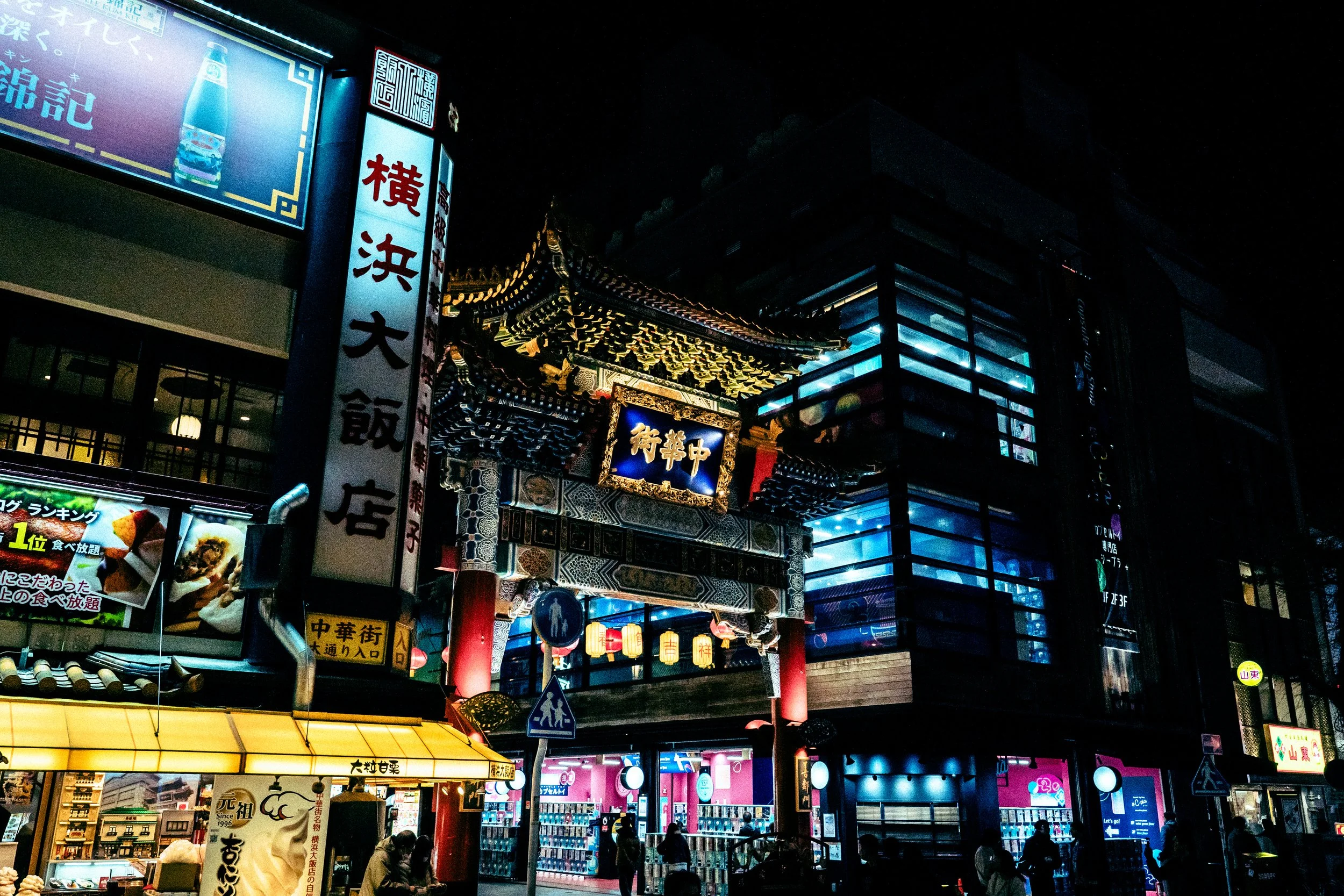 Night scene of an Asian street with illuminated signs, traditional architectural elements, and people walking below.