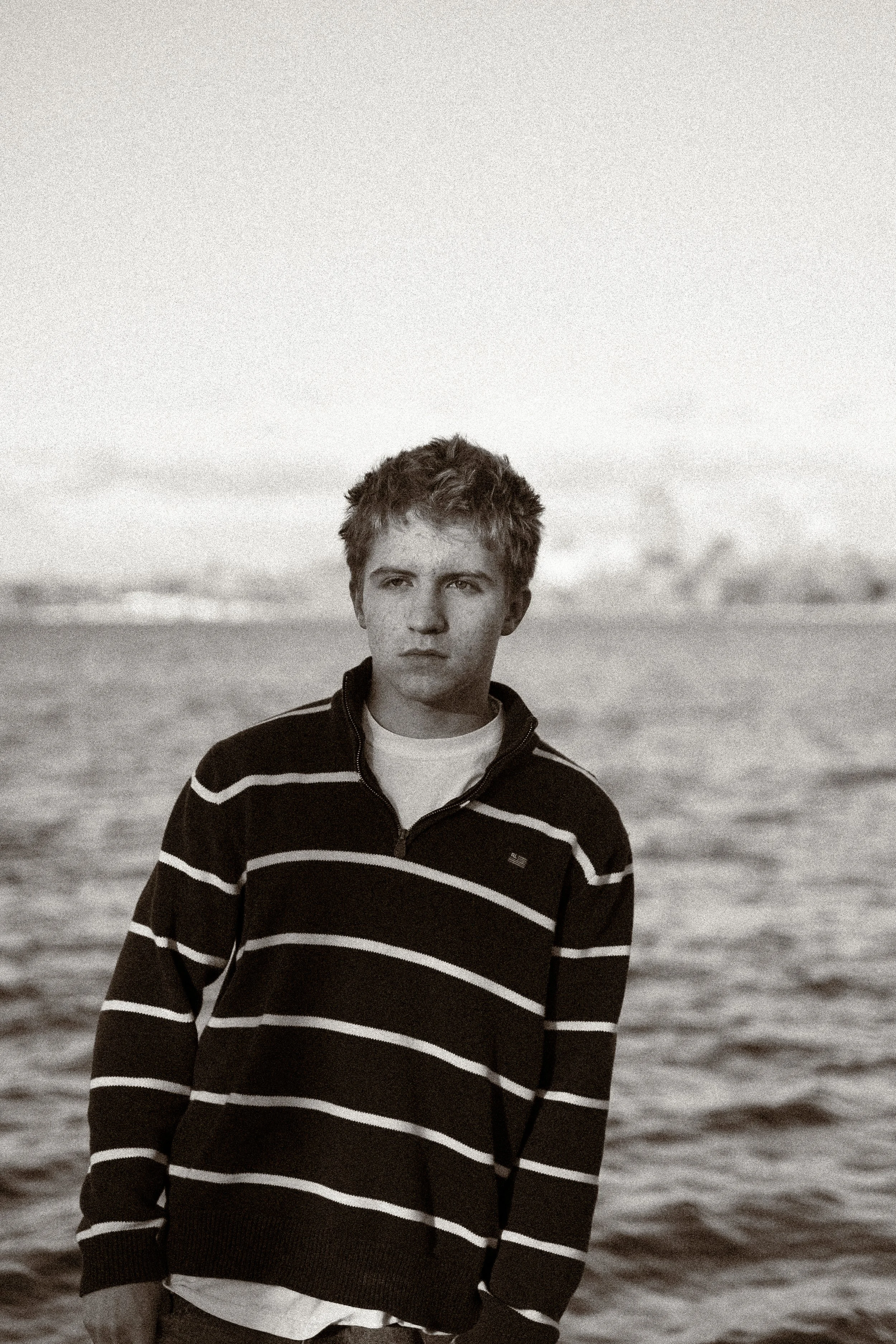A young man in a striped sweater standing outdoors near a body of water, with a city skyline in the background.
