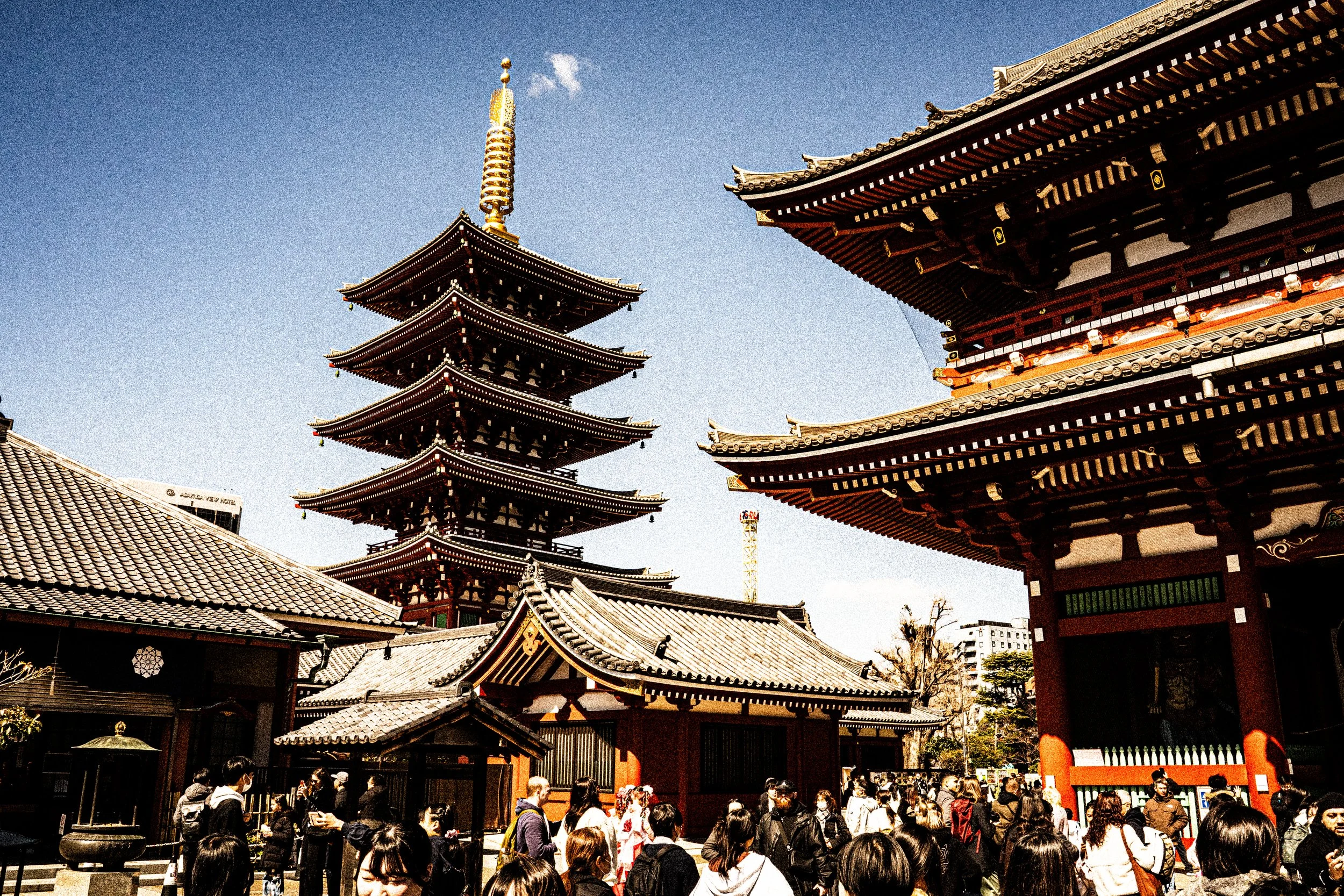 A traditional Japanese pagoda with multiple tiers and ornate eaves, surrounded by temple buildings and a crowd of people visiting the site.