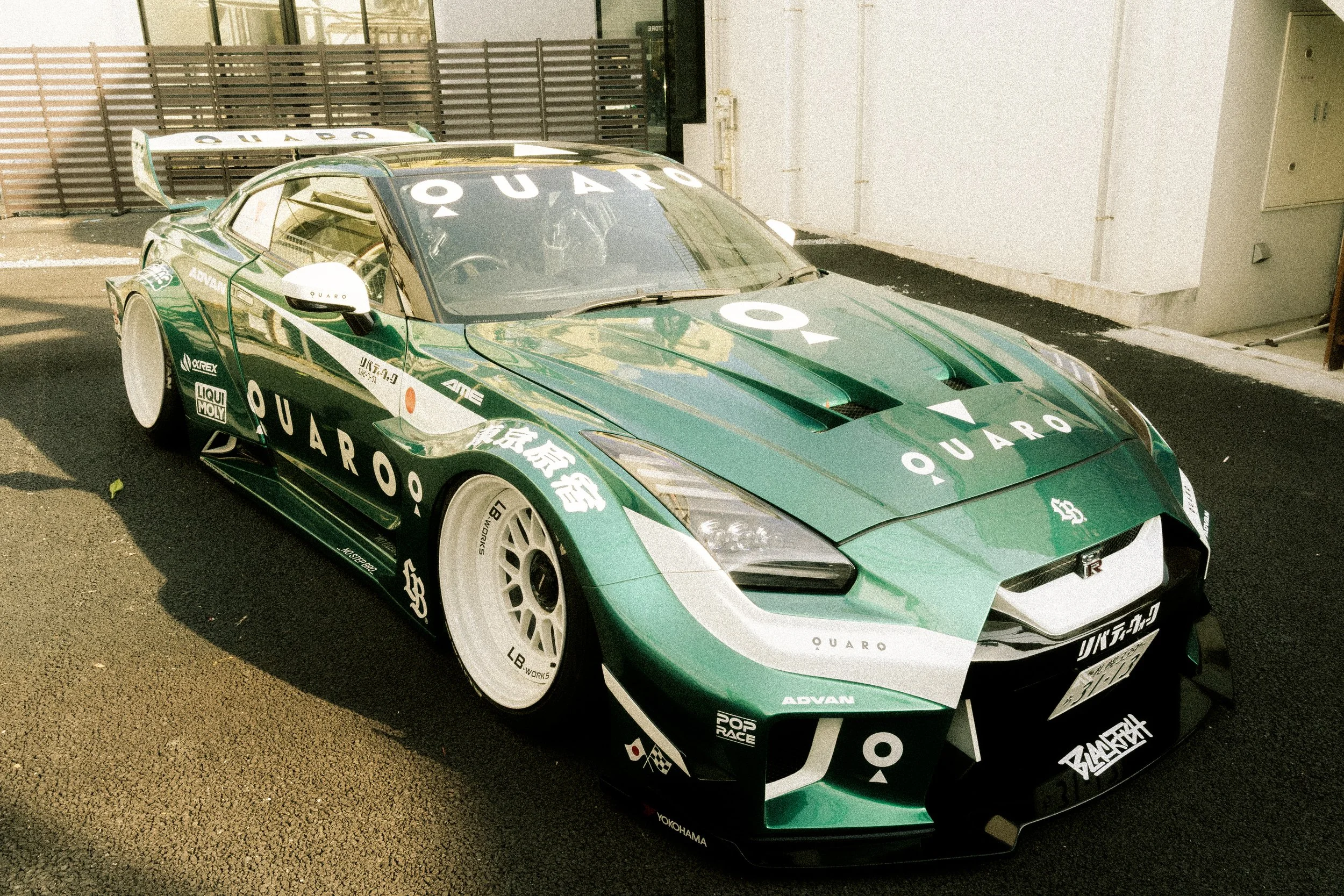A green race car with various sponsor logos and decals, including 'QUARO,' parked outdoors near a building and a wooden fence, with a rear wing and aerodynamic design.