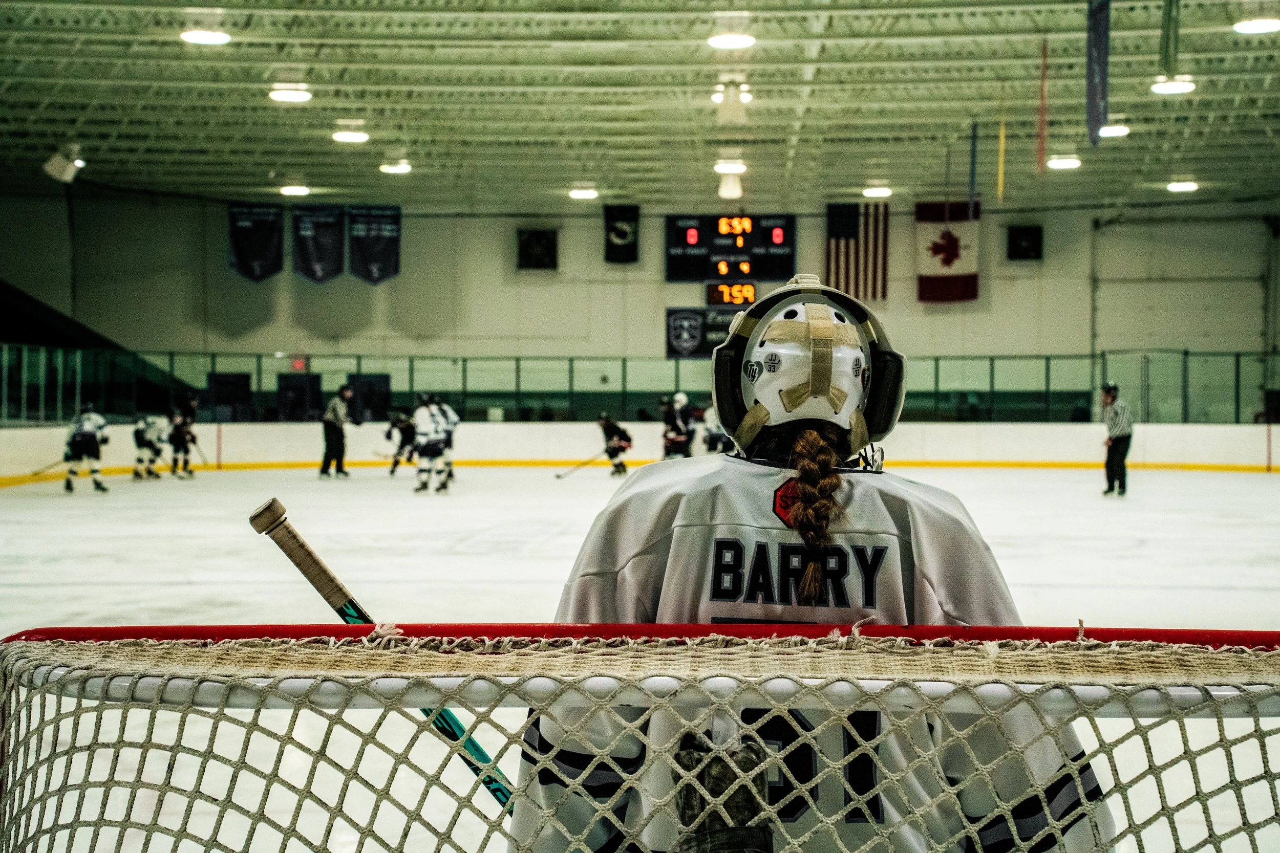 A hockey player with a braid and jersey that says 'BARRY' stands behind the goal net, watching a game on the ice rink with other players and referees.