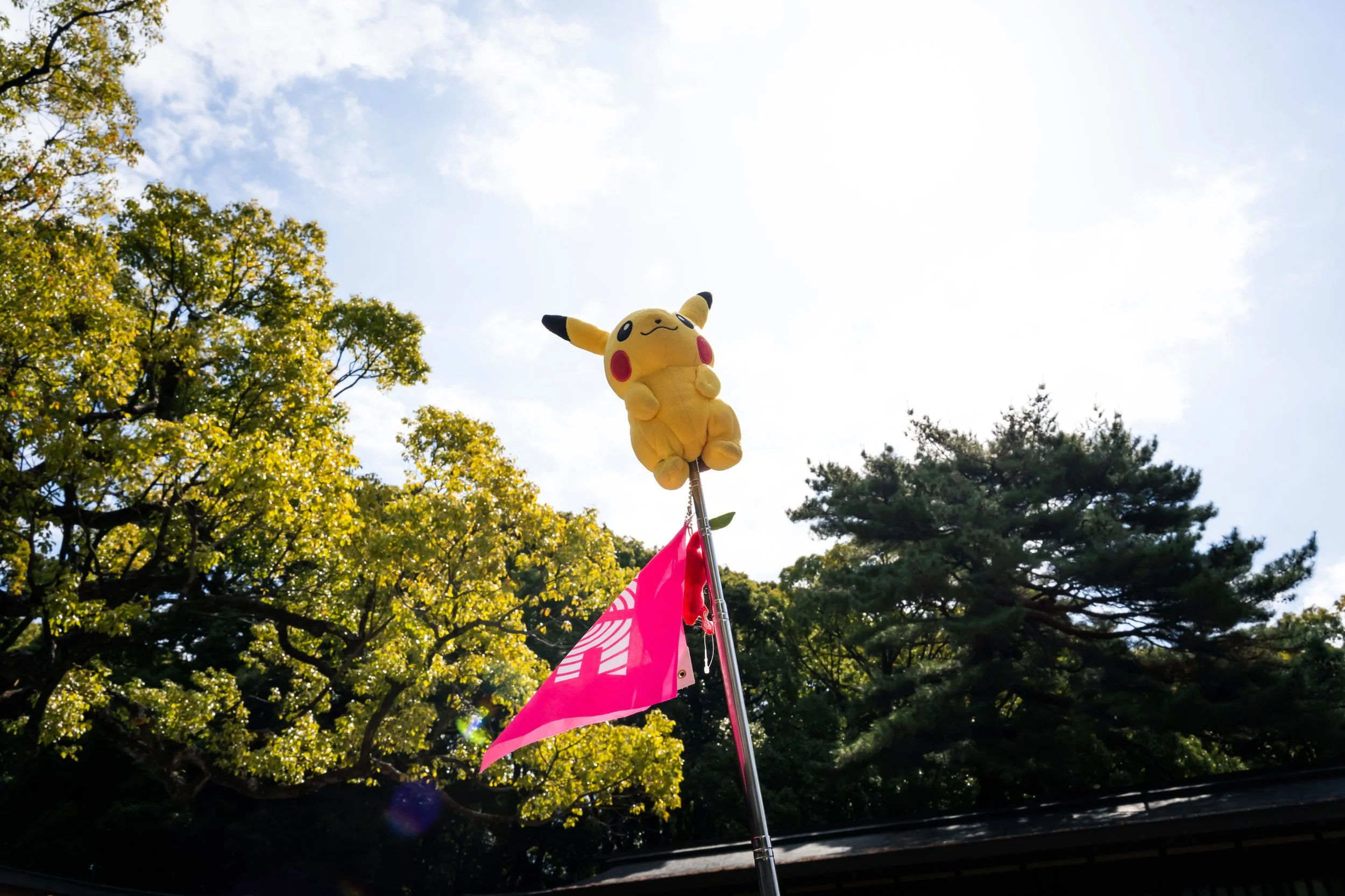 A Pikachu plush toy perched on a flagpole with pink and white flags, set against a backdrop of trees with green leaves and a partly cloudy sky.