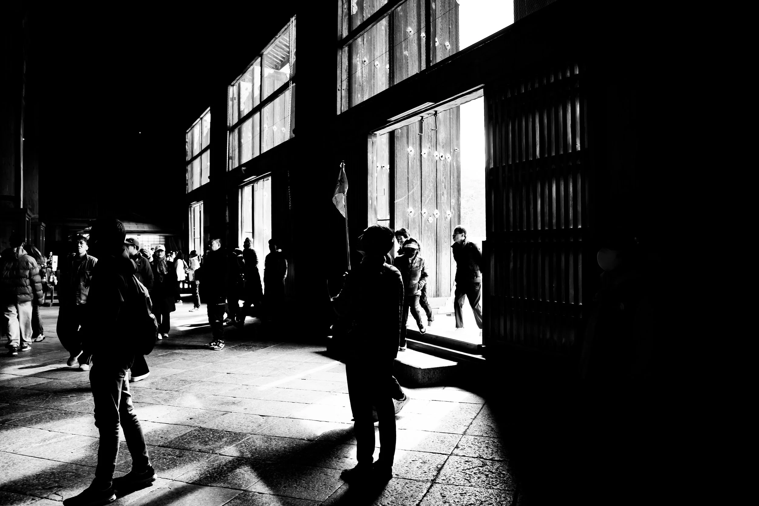 People walking inside a building with large open doors, some sunlight streaming through, creating silhouettes of the individuals.