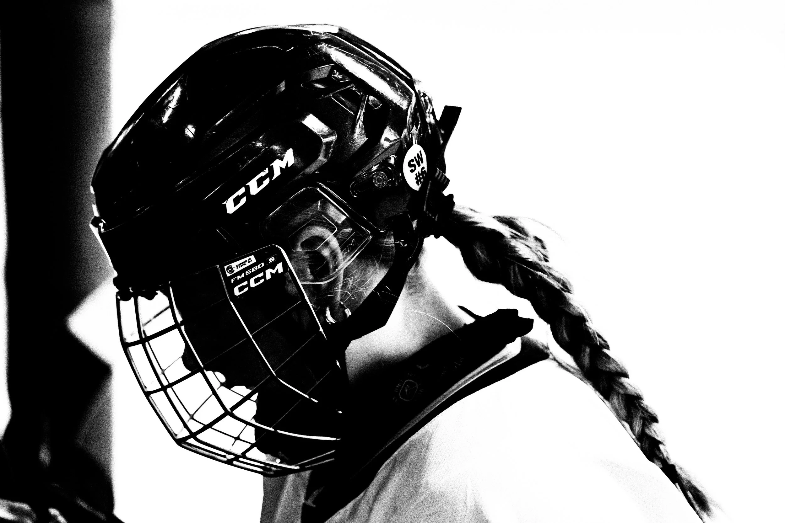 Black and white close-up of a hockey goaltender in gear, including a helmet with a cage, showing branding such as CCM and Sher-Wood, with a shoulder pad visible, against a plain background.