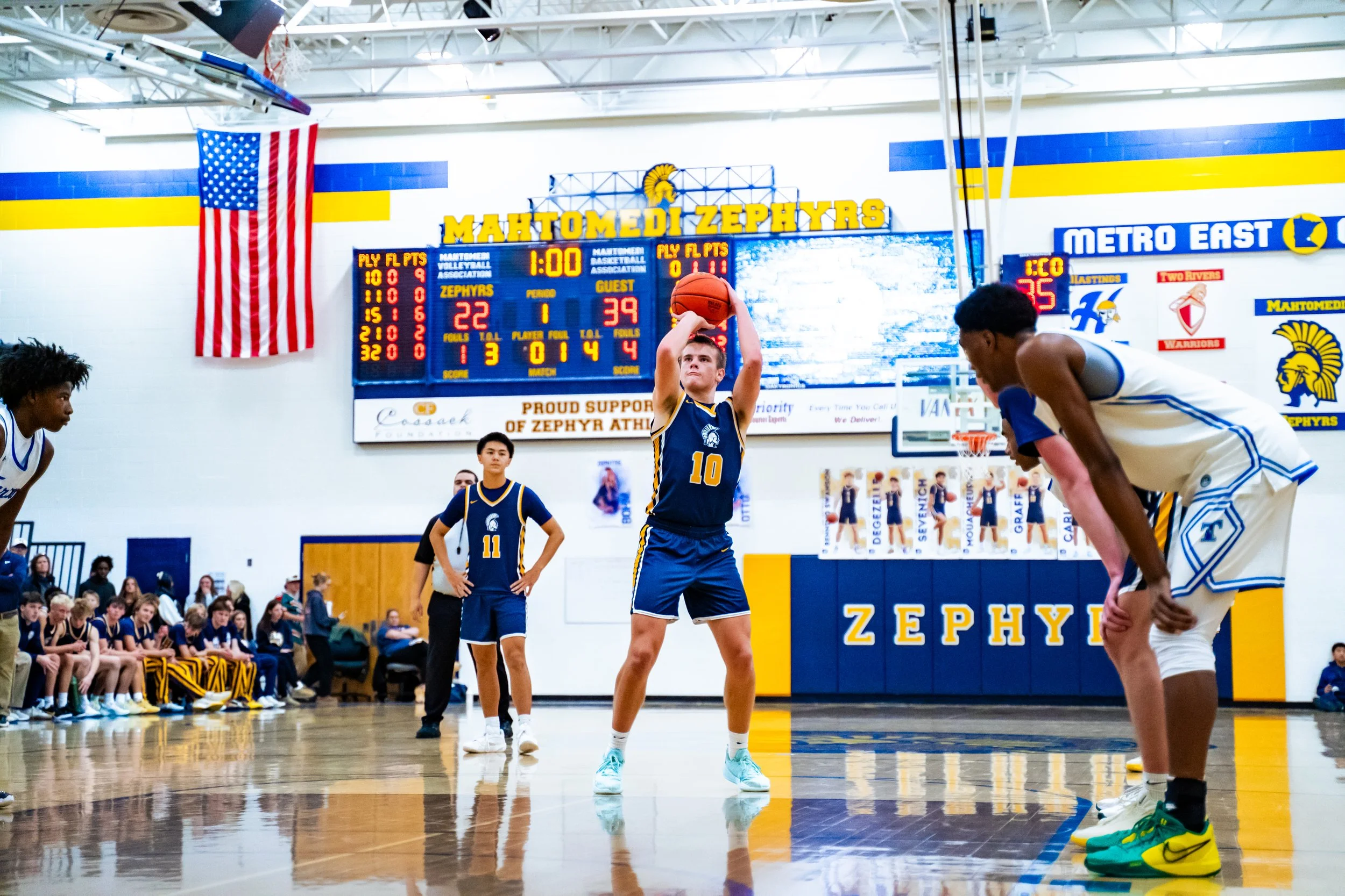 A young female basketball player in a blue and yellow uniform practices free throws during a game at Mahtomed Zephyrs gymnasium. She is holding a basketball above her head, preparing to shoot, while other players and spectators watch.
