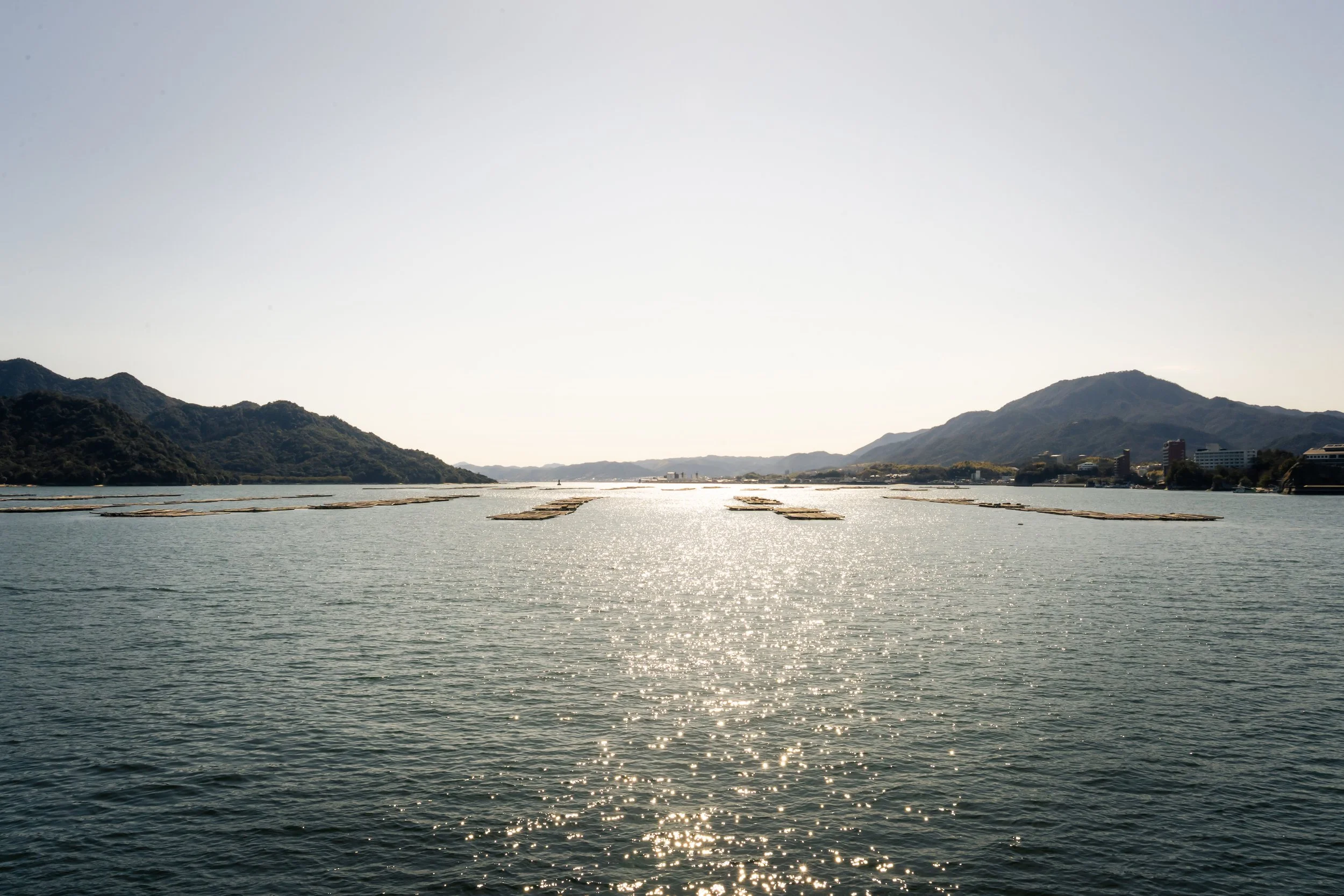 A body of water with floating platforms or nets, surrounded by green hills and mountains under a clear sky with the sun reflecting on the water