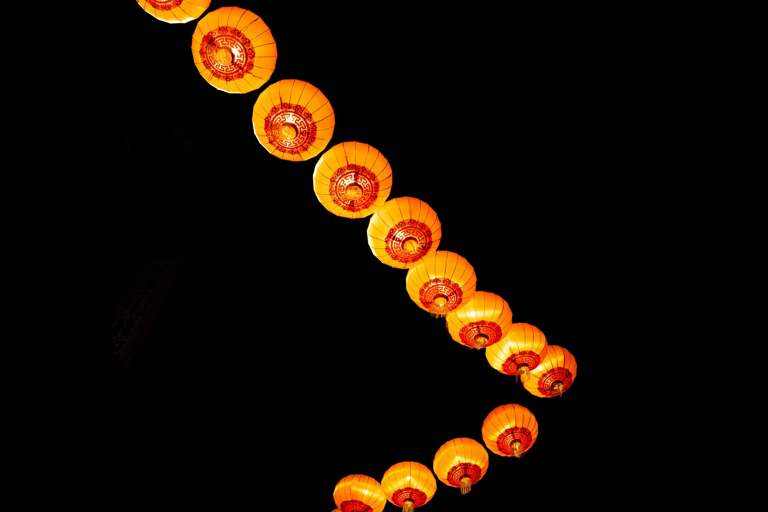 A string of illuminated orange paper lanterns with red decorative designs hanging in a row against a black night sky.
