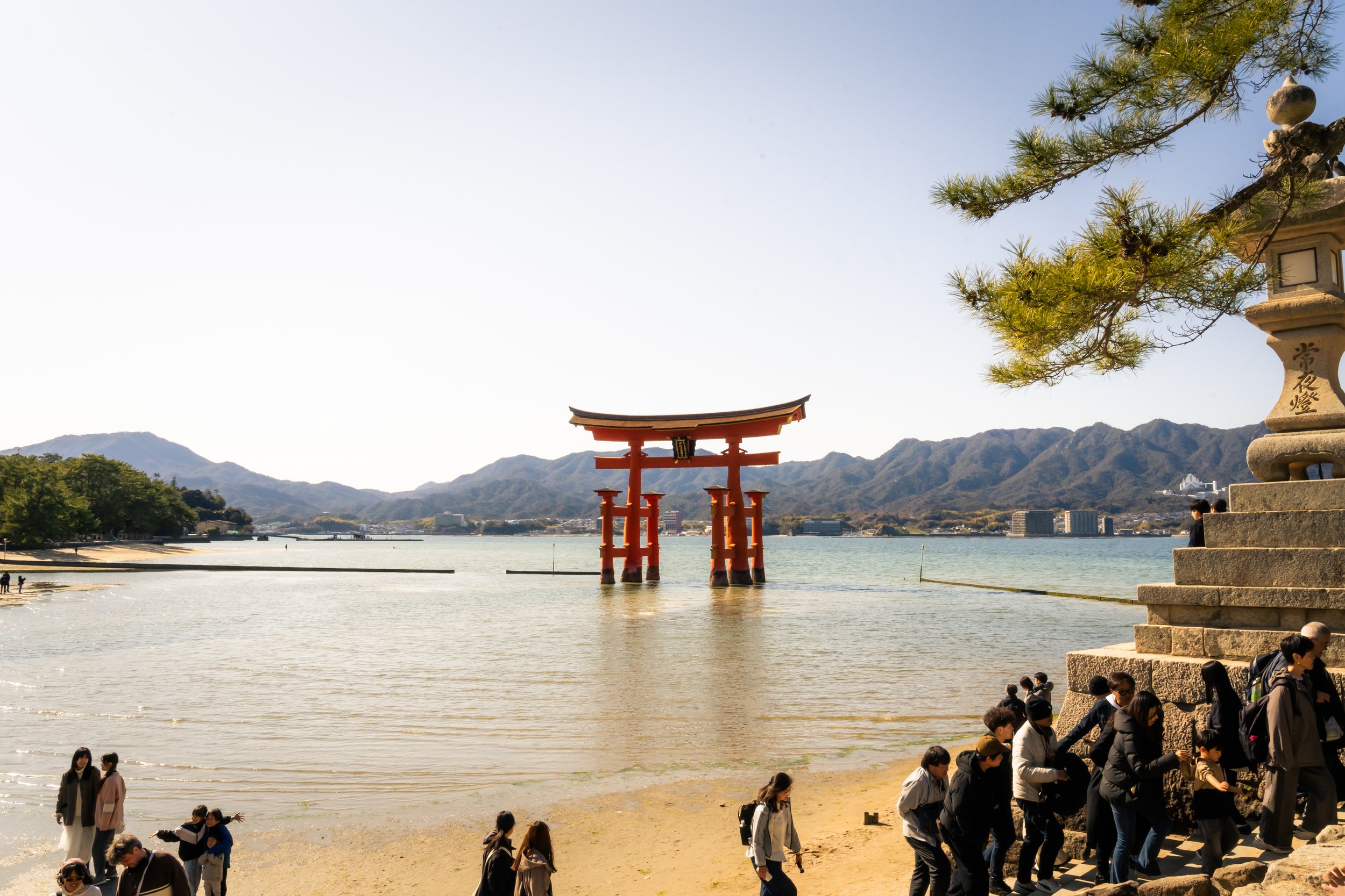People walking along a shoreline near a traditional Japanese torii gate standing in water, with mountain ranges in the background and a clear sky.
