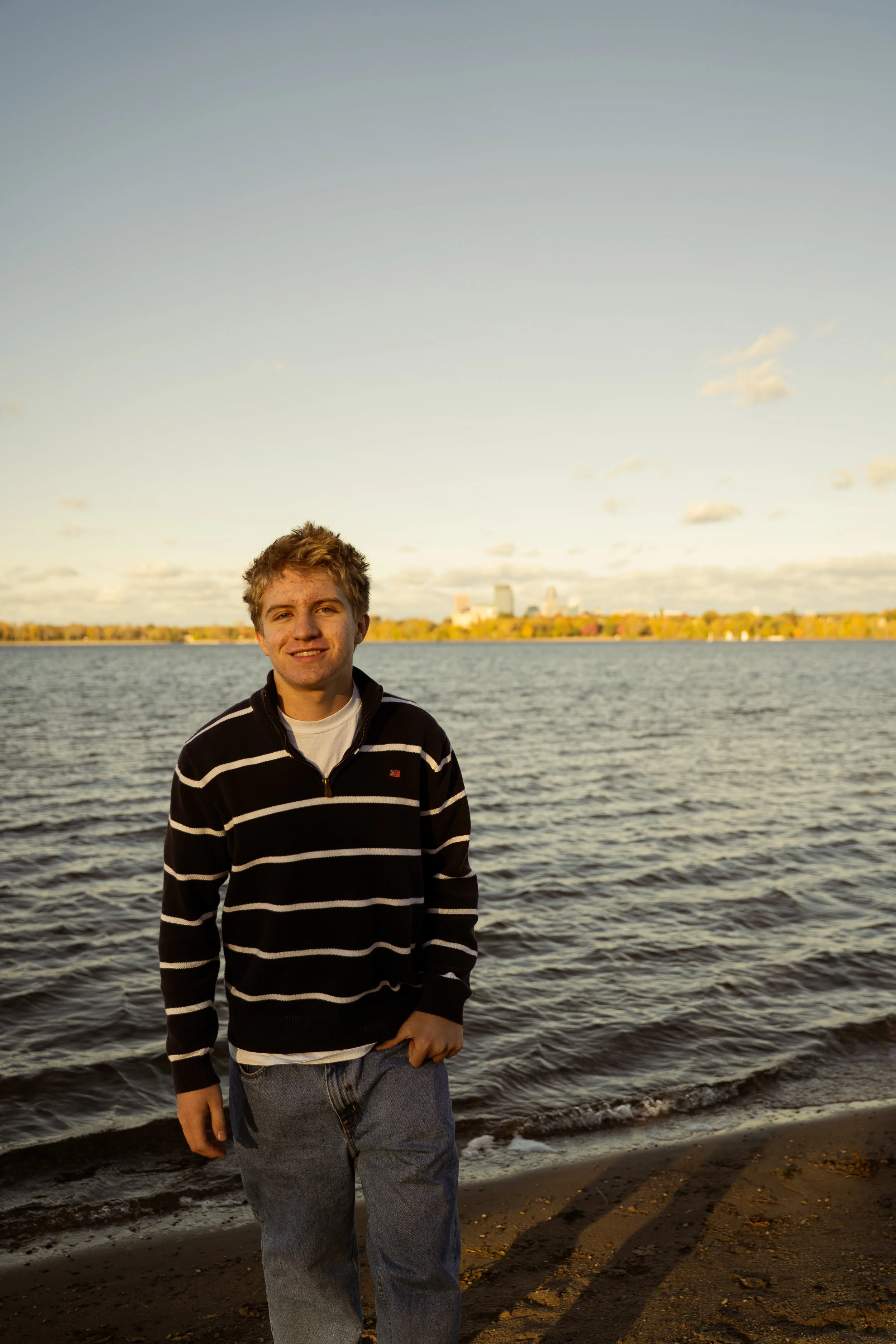 A young man with blond hair stands on a beach near the water, smiling, with city skyline in the background during sunset.