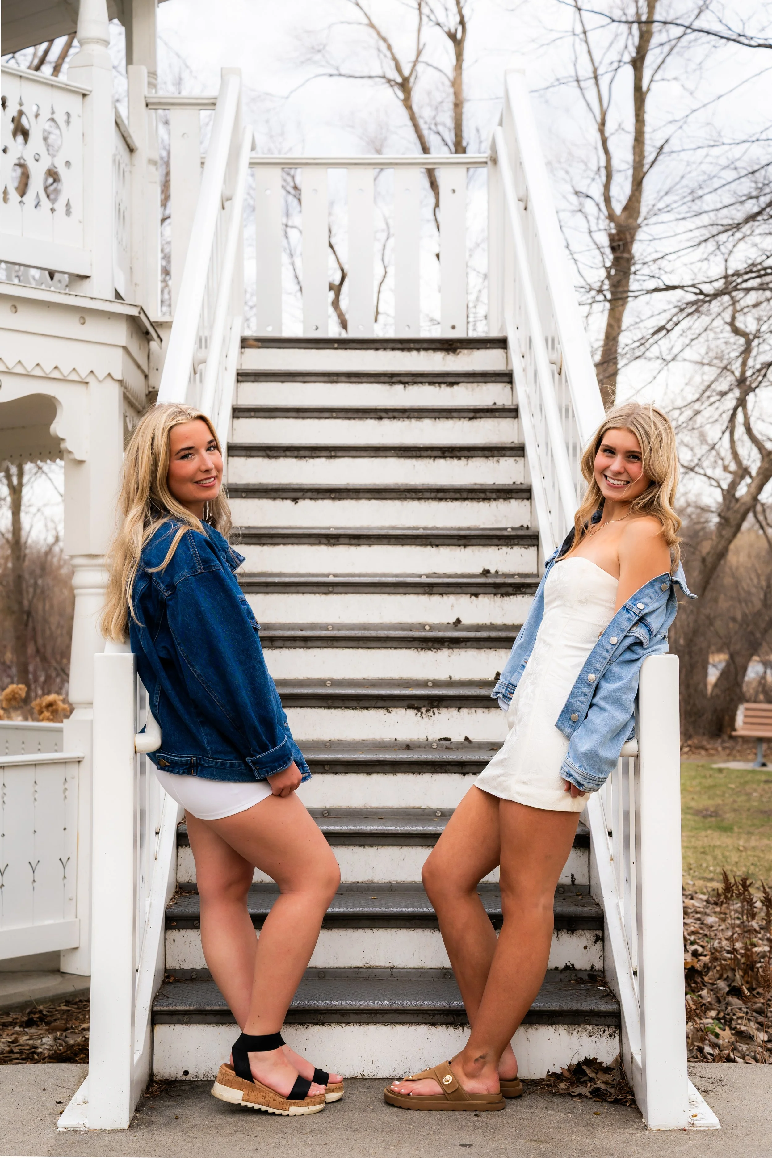 Two women with long blonde hair wearing denim jackets and dresses, posing on the stairs of a white porch with trees in the background.