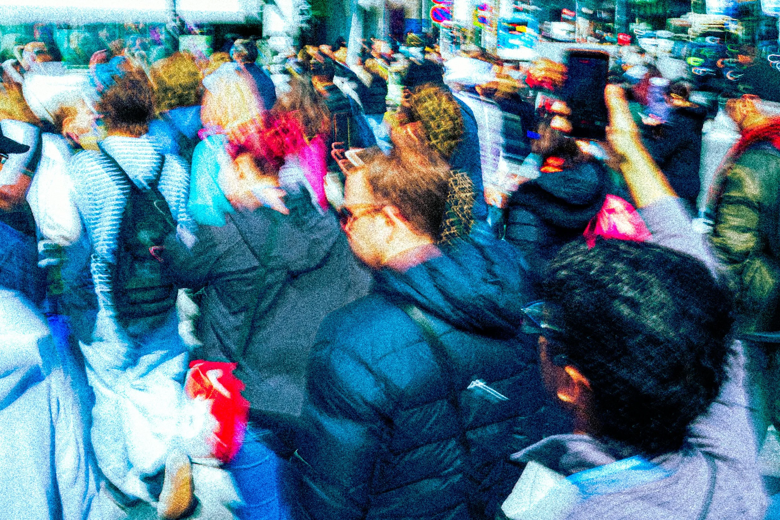 A crowd of people sitting closely together at a crowded indoor event, some holding phones, with colorful bags and clothing visible.