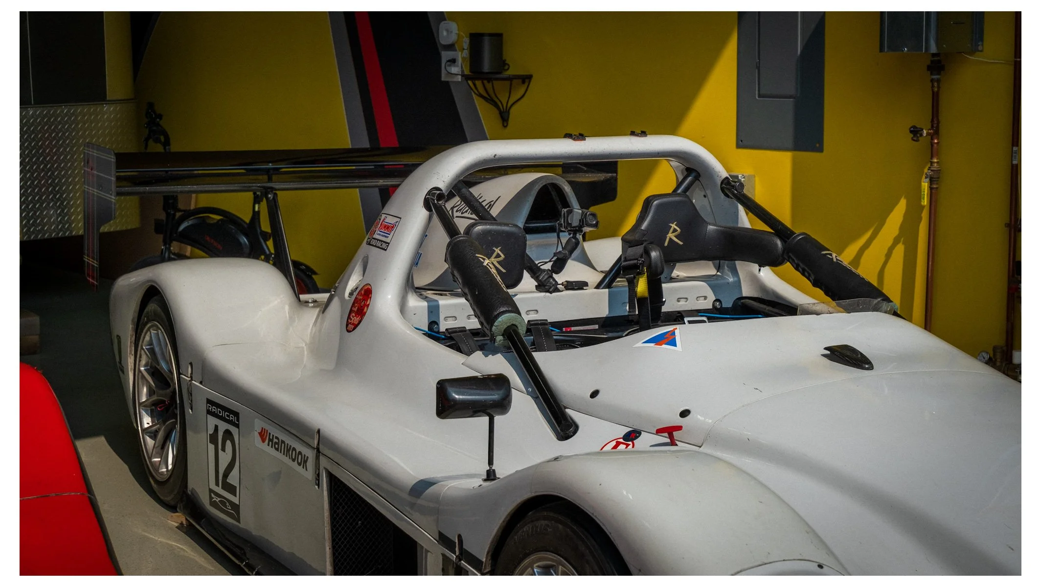 White race car with a large rear wing, racing number 12, and racing stickers inside a garage with yellow walls.