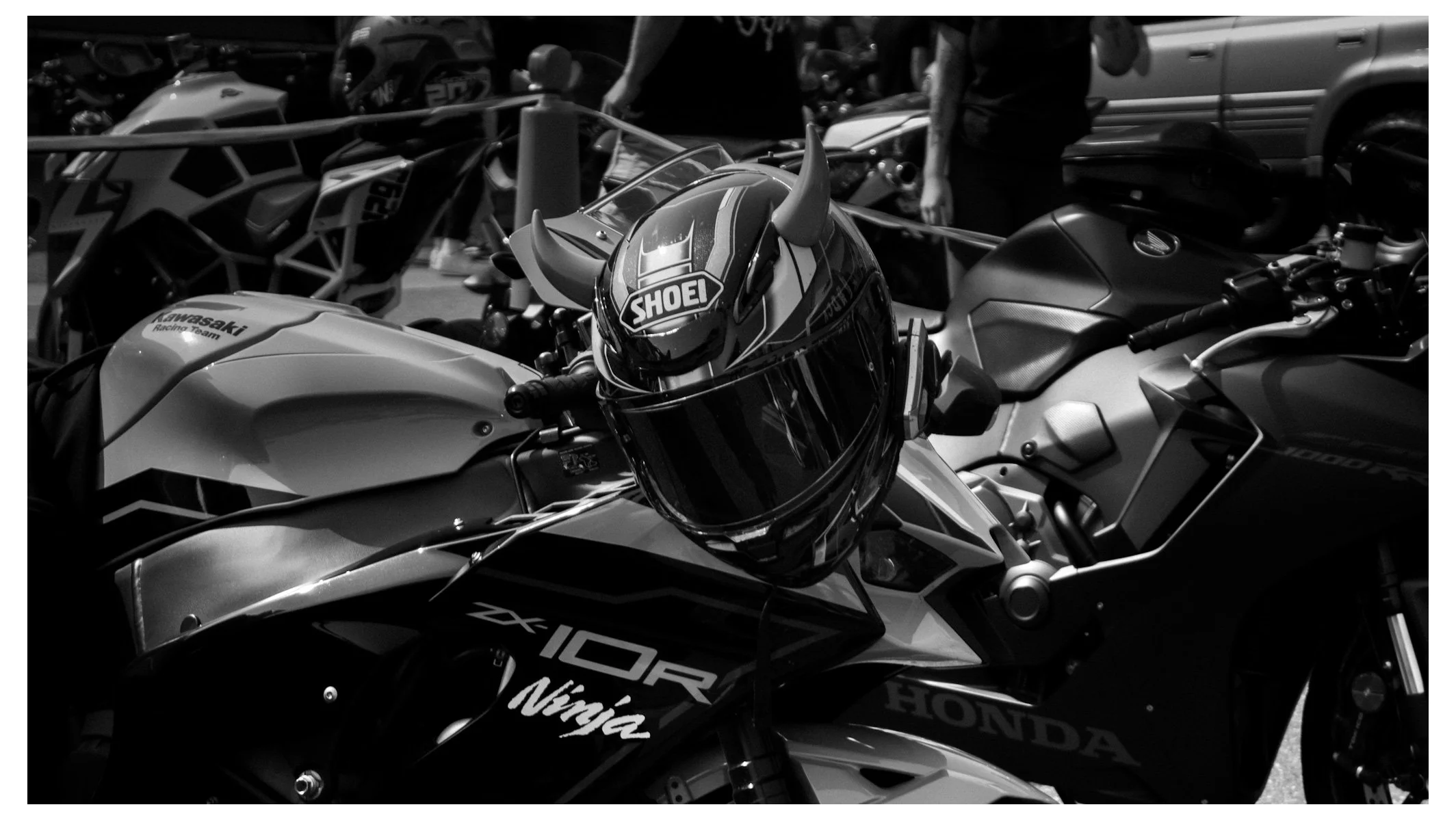 Black and white photo of several motorcycles parked closely together, with a focus on a Kawasaki Ninja motorcycle featuring a Shoei helmet on its handlebar and a Honda motorcycle nearby.