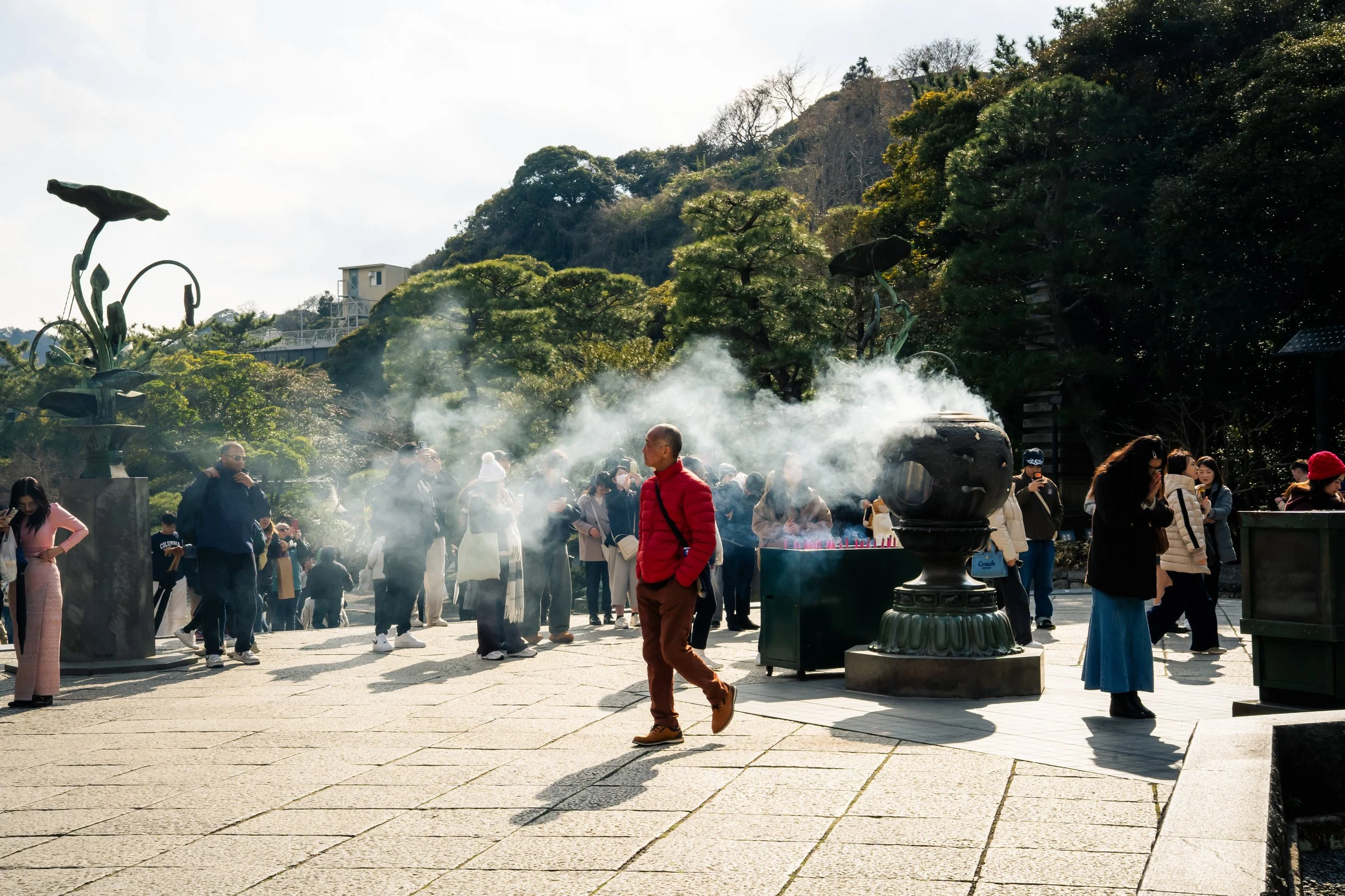People gathered around a steam-emitting incense burner at a traditional outdoor temple or shrine, with lush green trees and a hillside in the background.