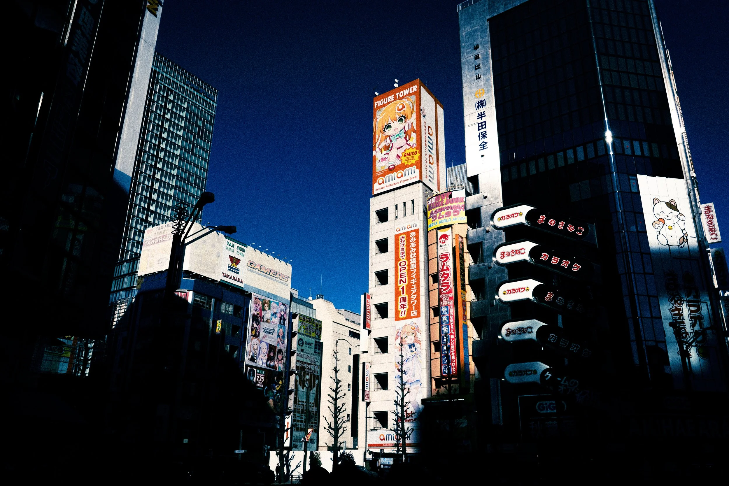 A cityscape with tall buildings featuring colorful advertisements and billboards, including one with an anime-style character, under a clear blue sky, with street-level trees and signs.