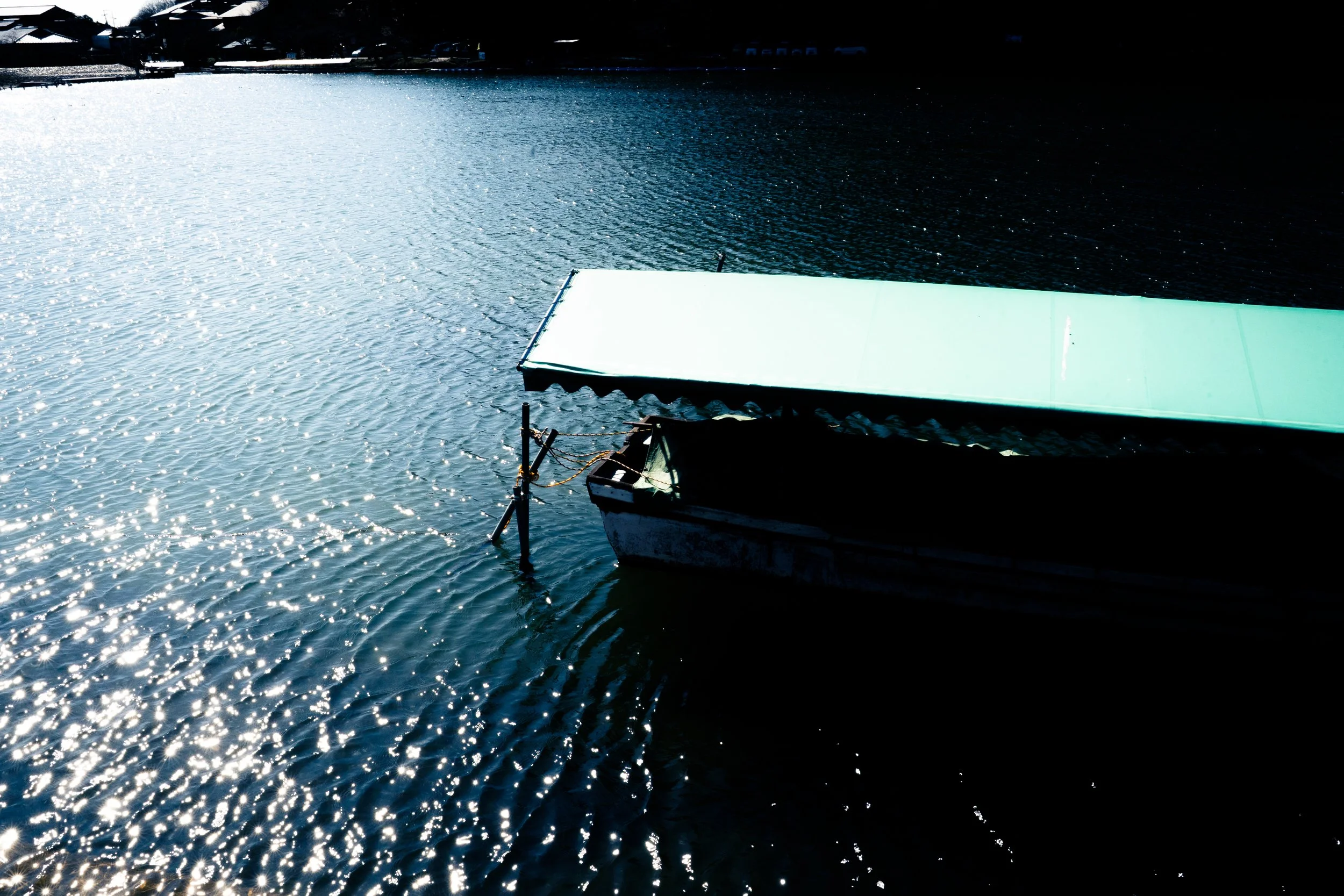 A dock with a boat attached, floating on a body of water with sunlight reflecting off the surface, and a dark background where the shoreline or trees are barely visible.