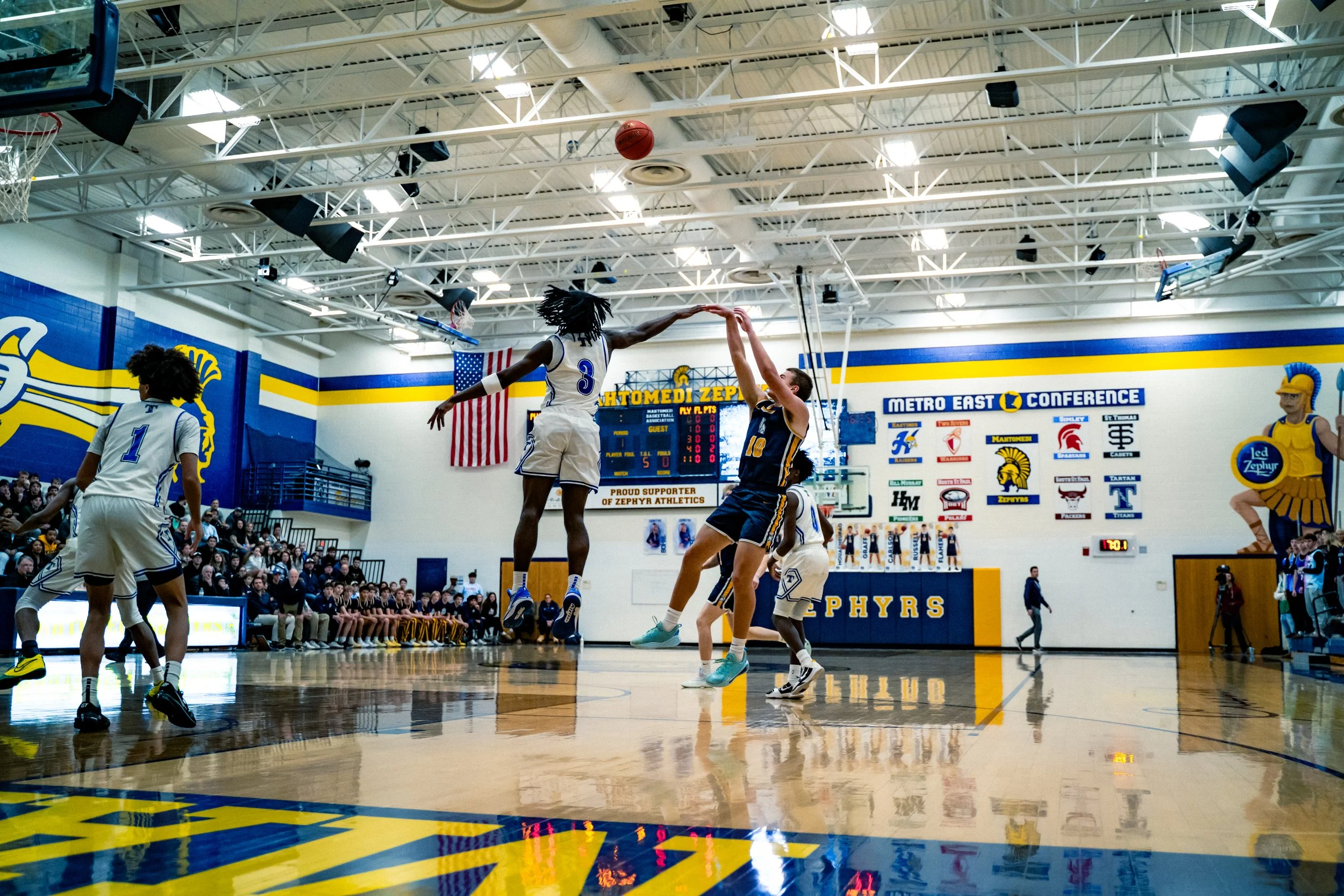 A basketball game in a gymnasium with players jumping to catch or contest a basketball near the hoop. Spectators are seated on the bleachers, and banners and scoreboards are visible on the walls.