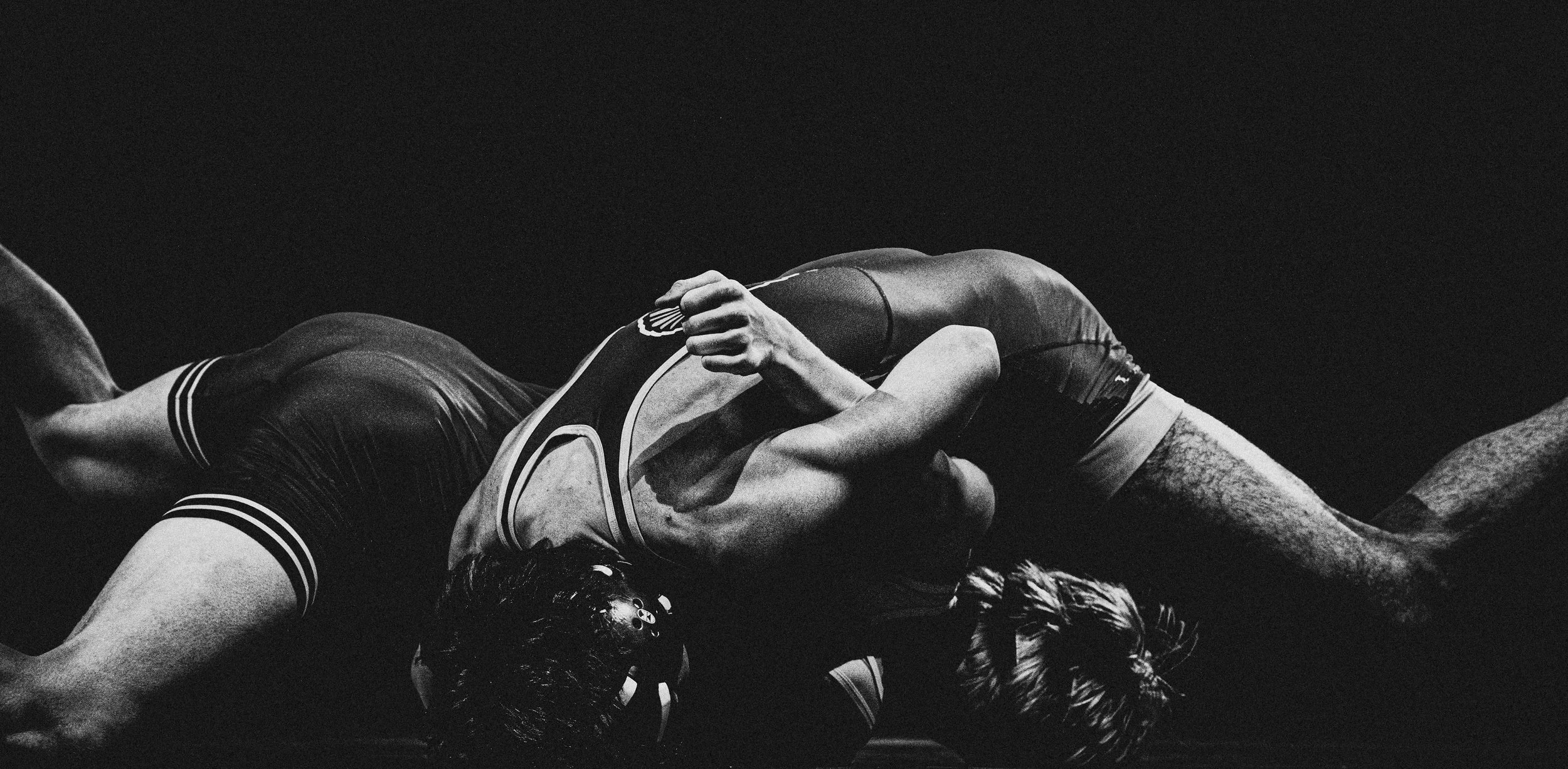 Two wrestlers engaged in a match, one lying on top of the other, in a black and white photograph.