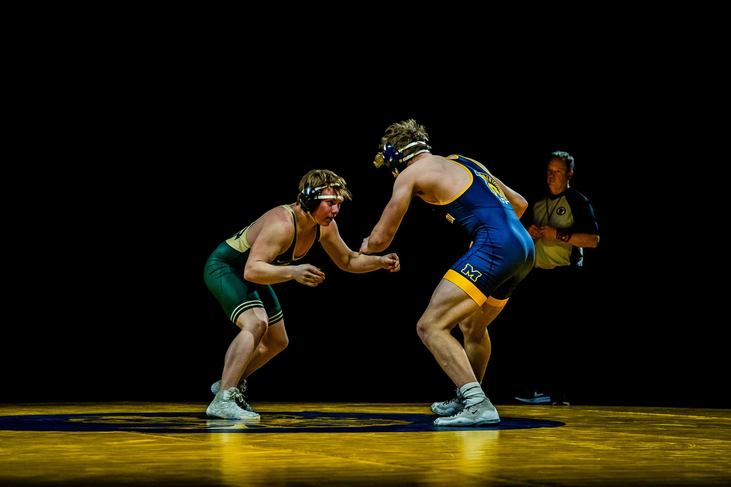 Two male wrestlers in a match, wearing headgear and singlets, engaged in a stance on a wrestling mat with a referee observing.