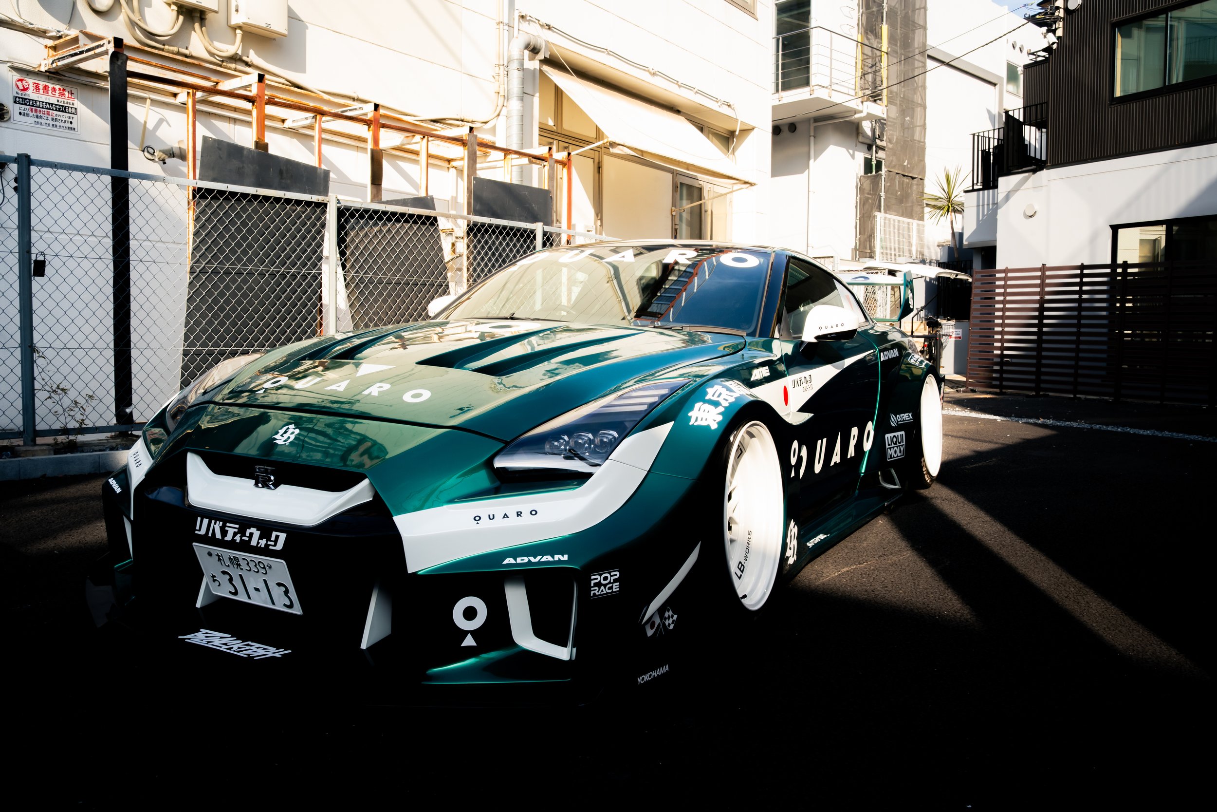 A green and white race car with various sponsorship stickers parked outdoors in an urban setting, with buildings and fences in the background.