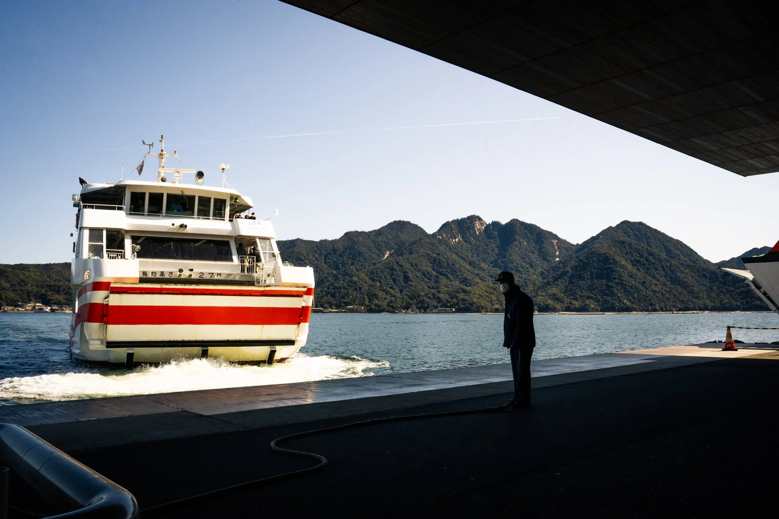 A ferry boat on the water with mountains in the background, viewed from under a bridge or dock, with a person wearing a face mask standing on the dock.