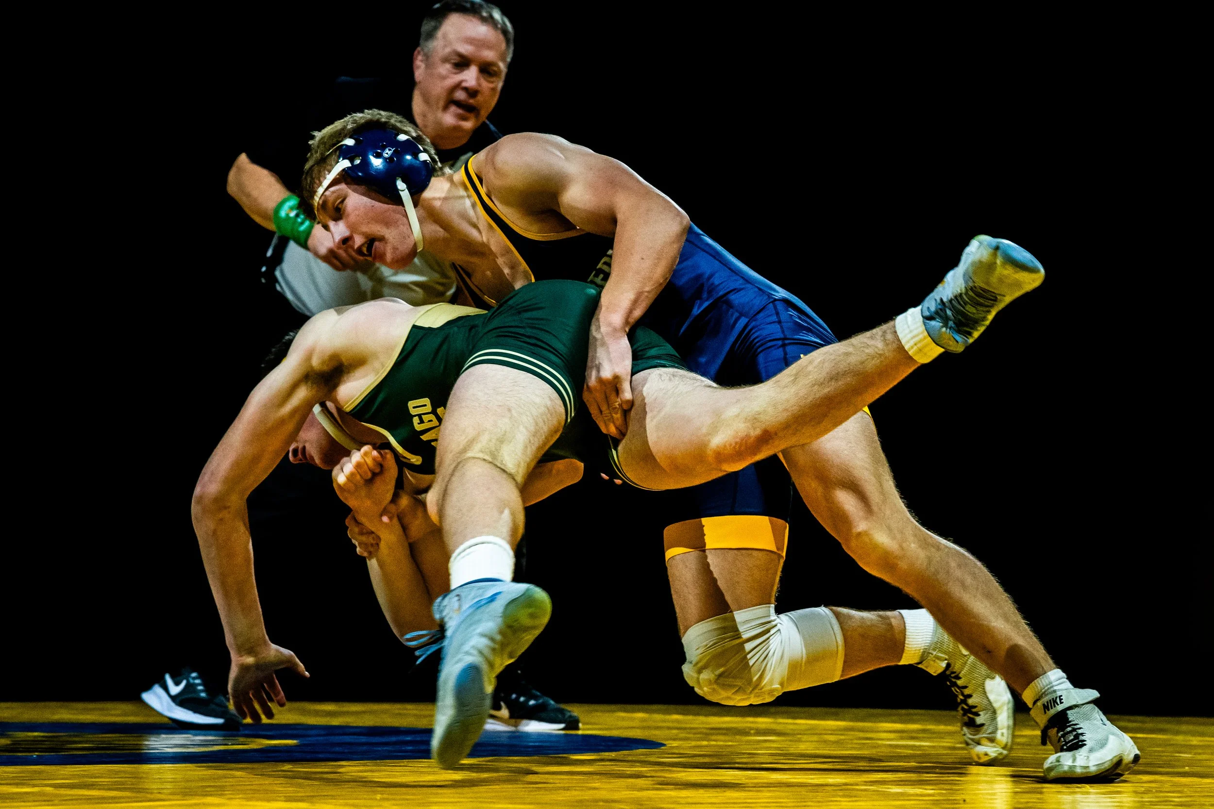 Two wrestlers compete on a wrestling mat with a referee watching closely in the background.