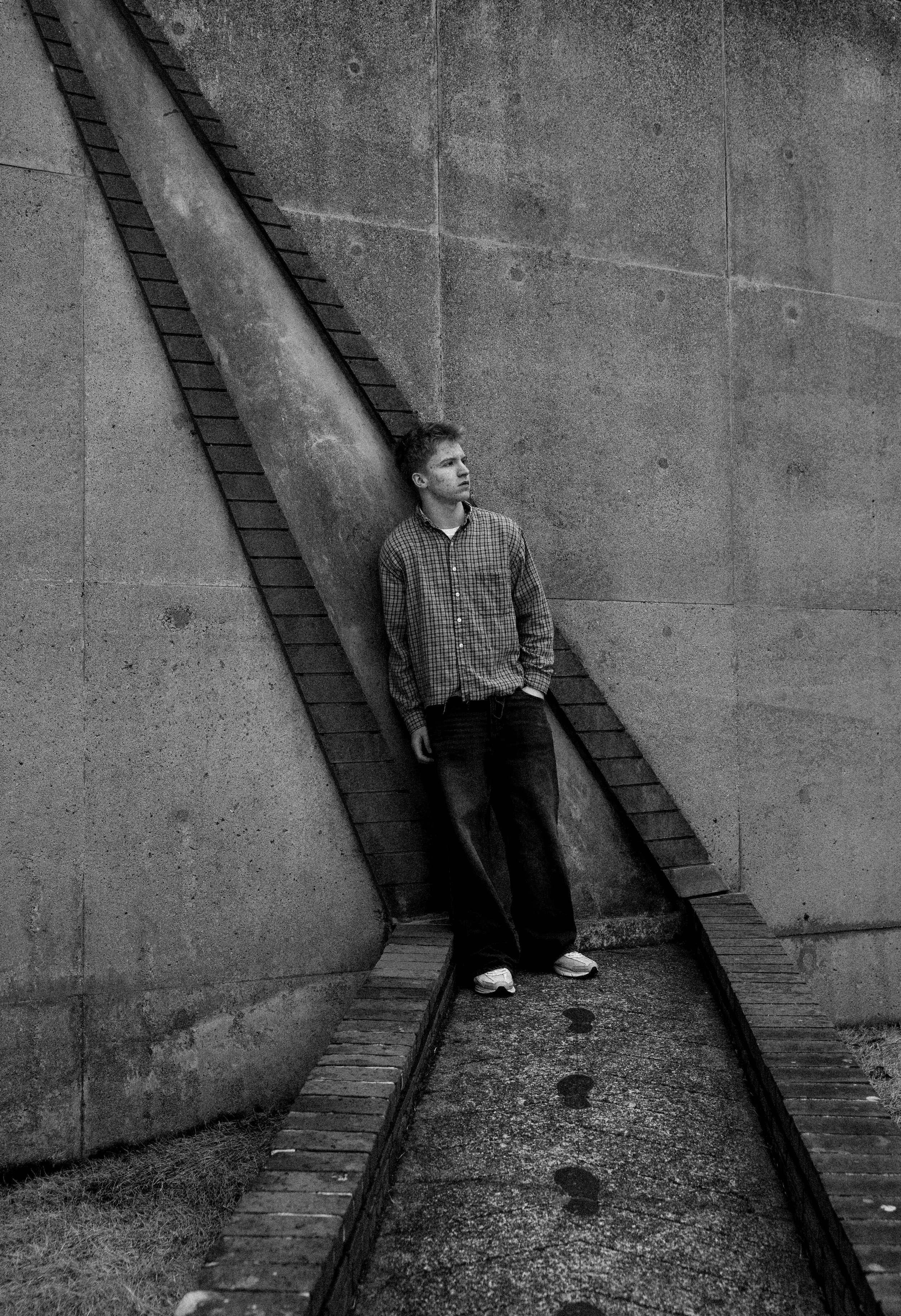 A young man wearing a checkered shirt, jeans, and sneakers stands against a large concrete wall, with his hands in his pockets and looking off to the side. The wall has stairs built into it, creating a unique geometric pattern.