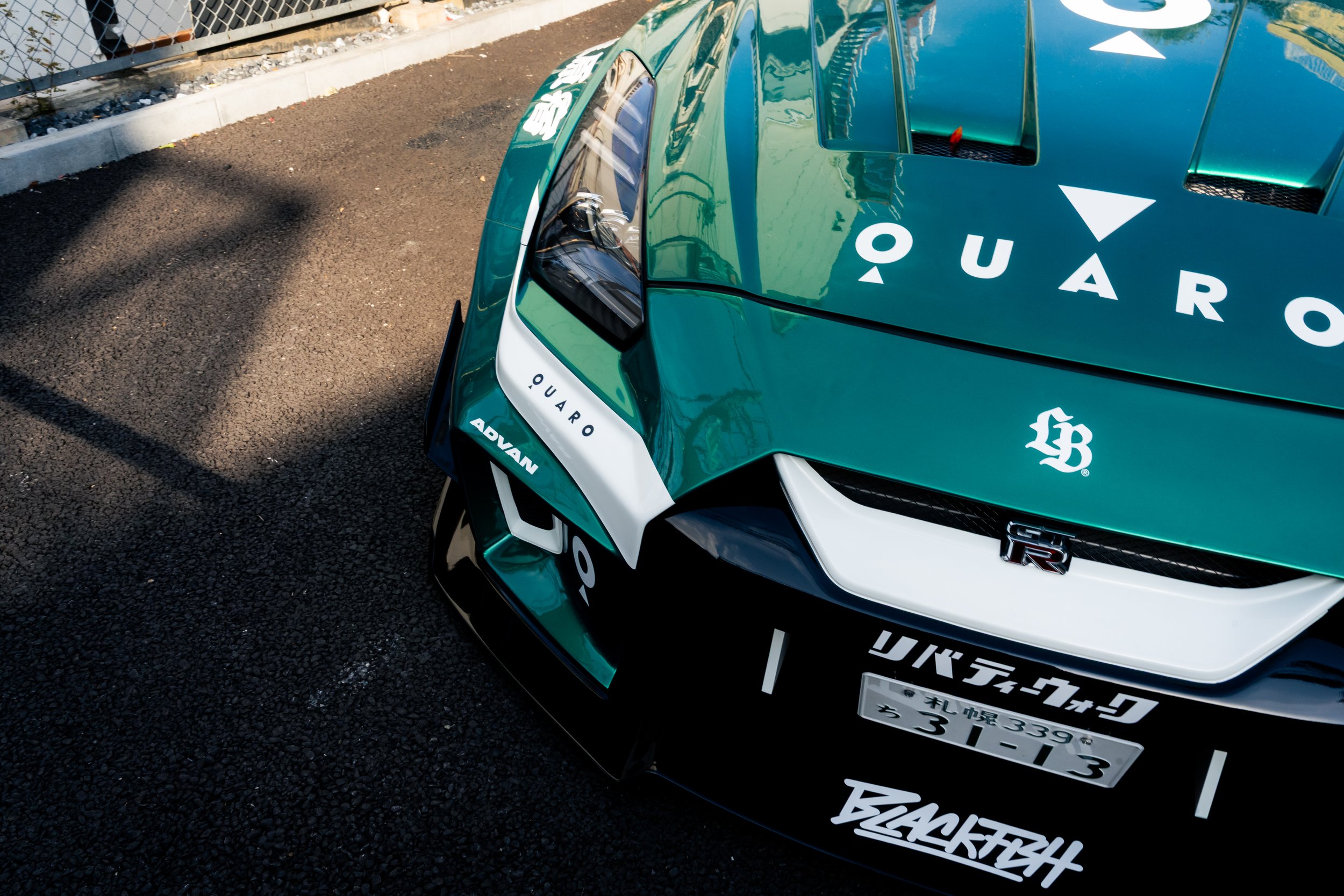 Close-up of a teal-colored race car with racing decals, Japanese license plate, and sponsorship logos, parked on asphalt with a chain-link fence and sidewalk in the background.