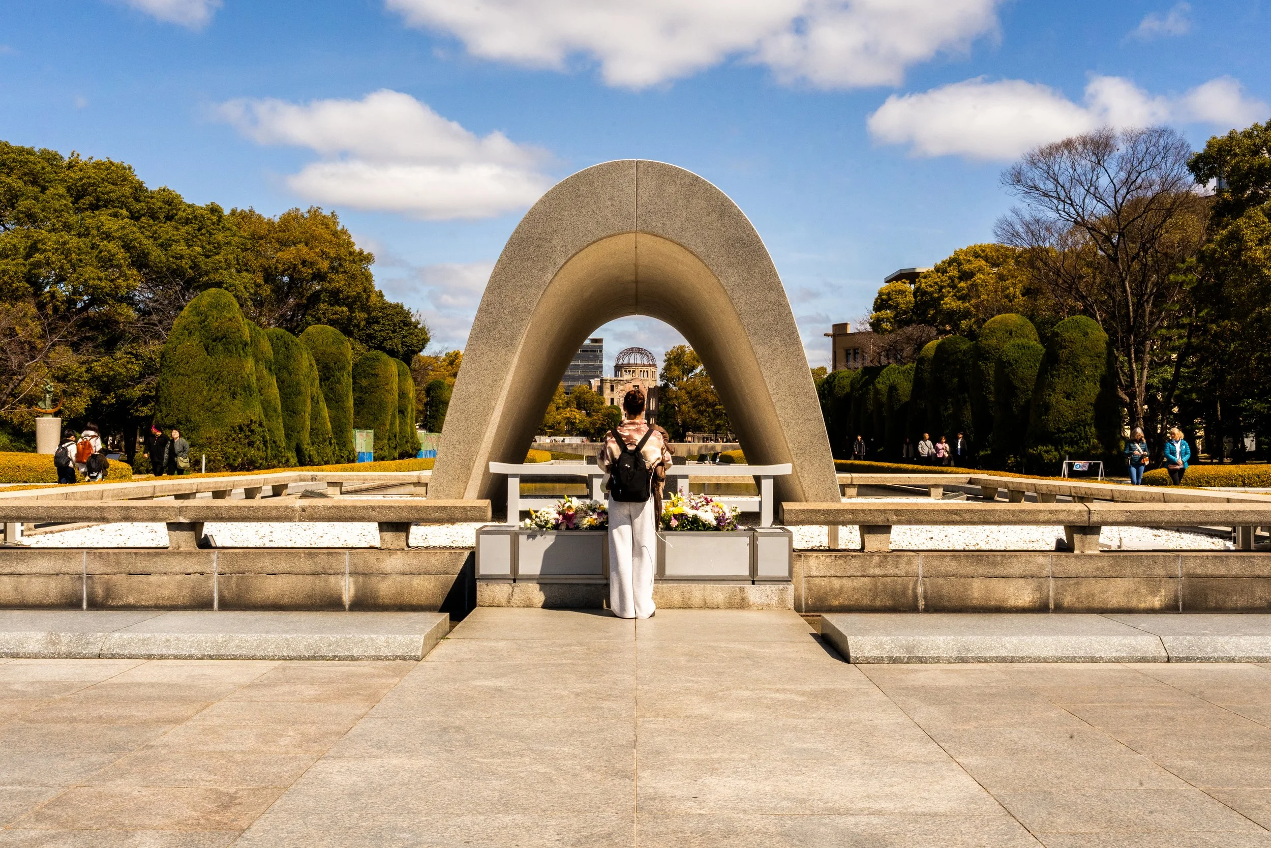 A woman standing in front of the Hiroshima Peace Memorial, also known as the Atomic Bomb Dome, with a flower arrangement nearby. The monument is a concrete arch and is situated in a park with trees and other visitors.