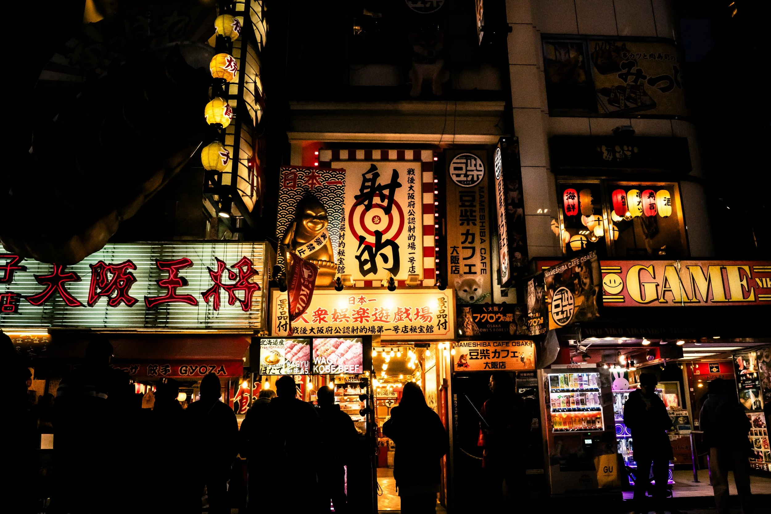 Night scene of a crowded street with illuminated signs and lanterns in Japanese, featuring shop fronts and silhouettes of people walking.