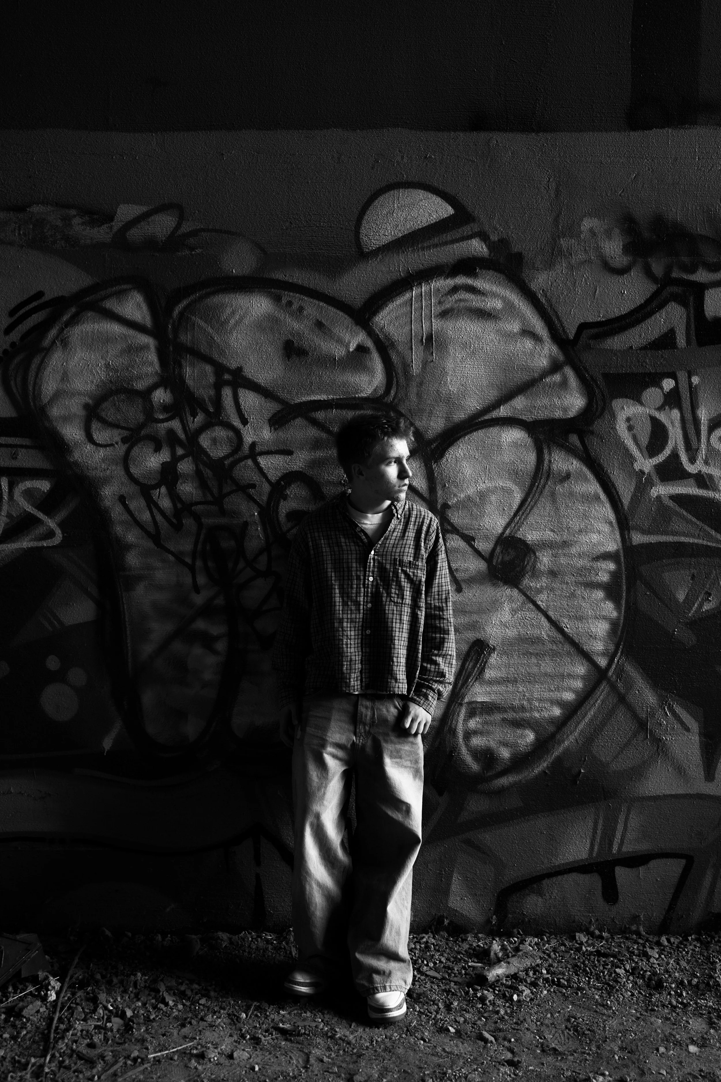 A young man in baggy pants and a checkered shirt stands against a graffiti-covered wall, looking to his right with a serious expression.