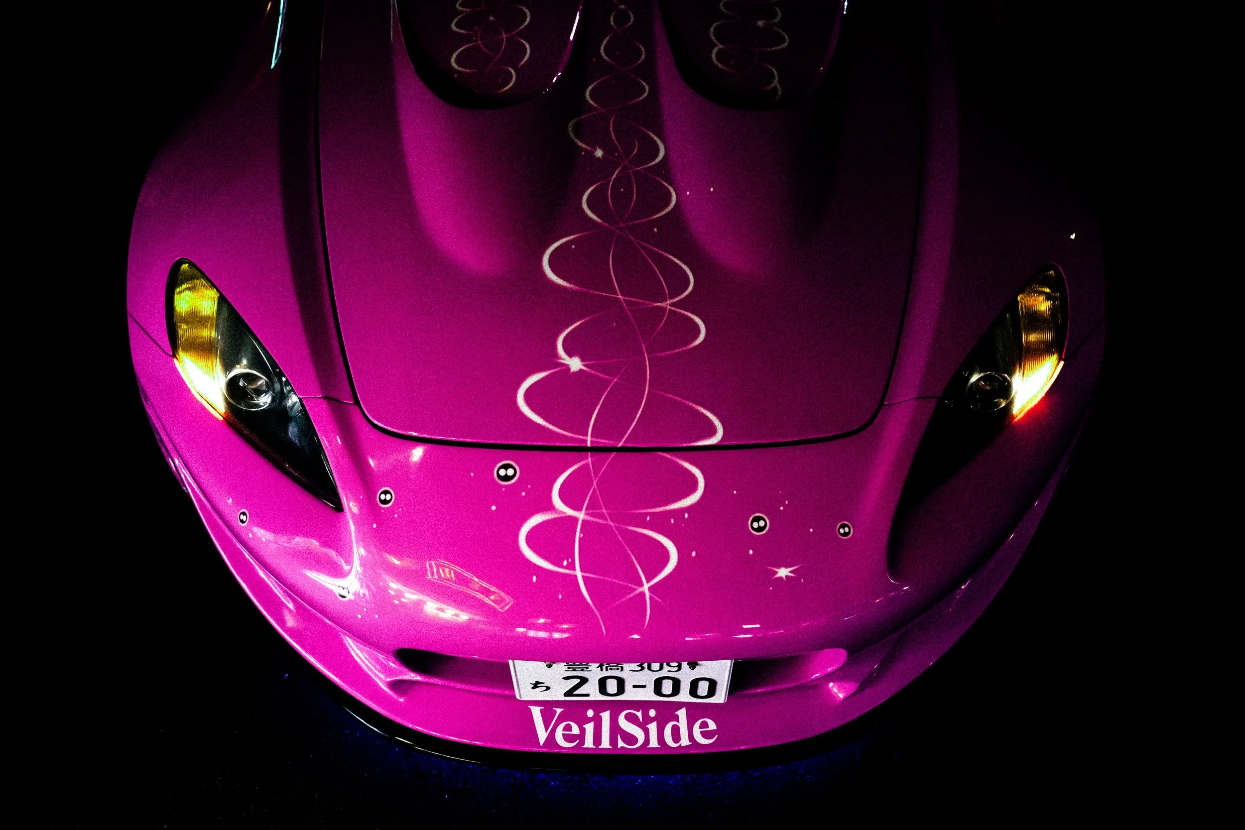 Close-up of a pink scooter with decorative white swirl patterns and small black and white yin-yang symbols, showing the front section, headlight, and a license plate.
