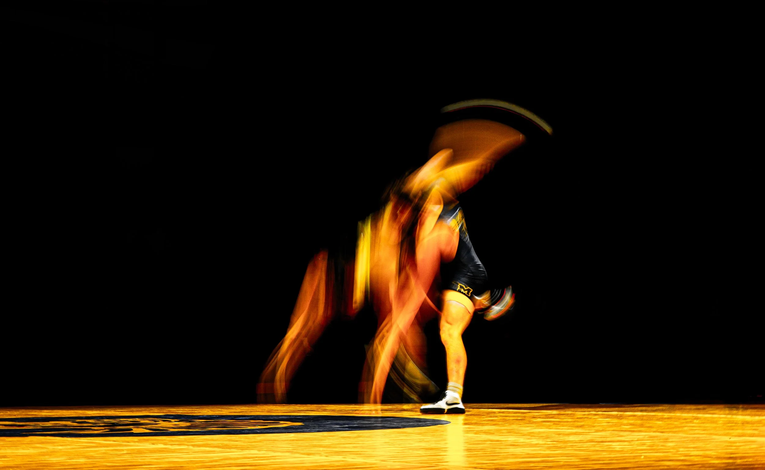 A person in a yellow and black sports jersey and white shoes is captured mid-action, possibly during a dance or sports event, against a dark background.