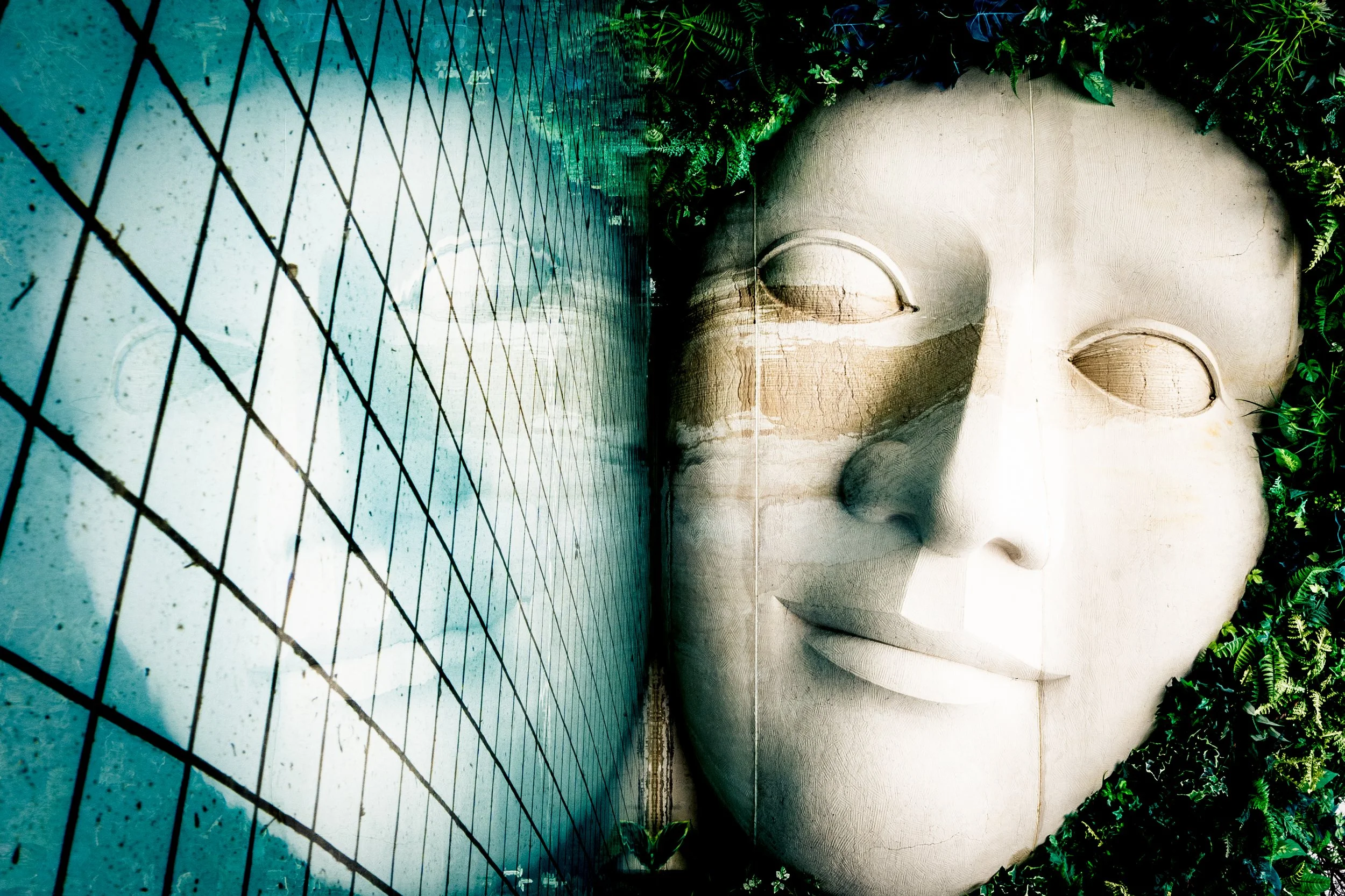 A white sculpted face relief on a wall surrounded by green plants and foliage, with a tiled area in the foreground.