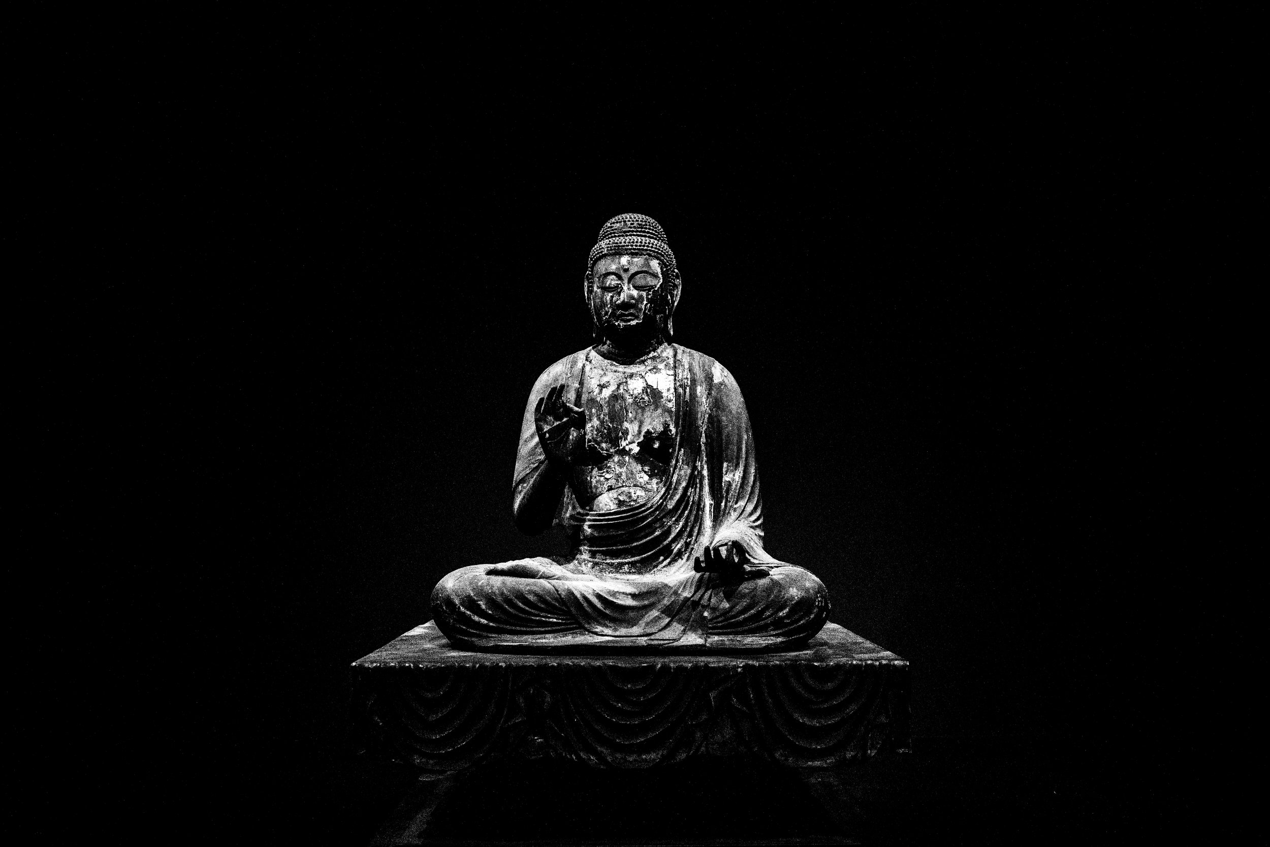 A black and white photograph of an ancient, weathered seated Buddha statue against a dark background.