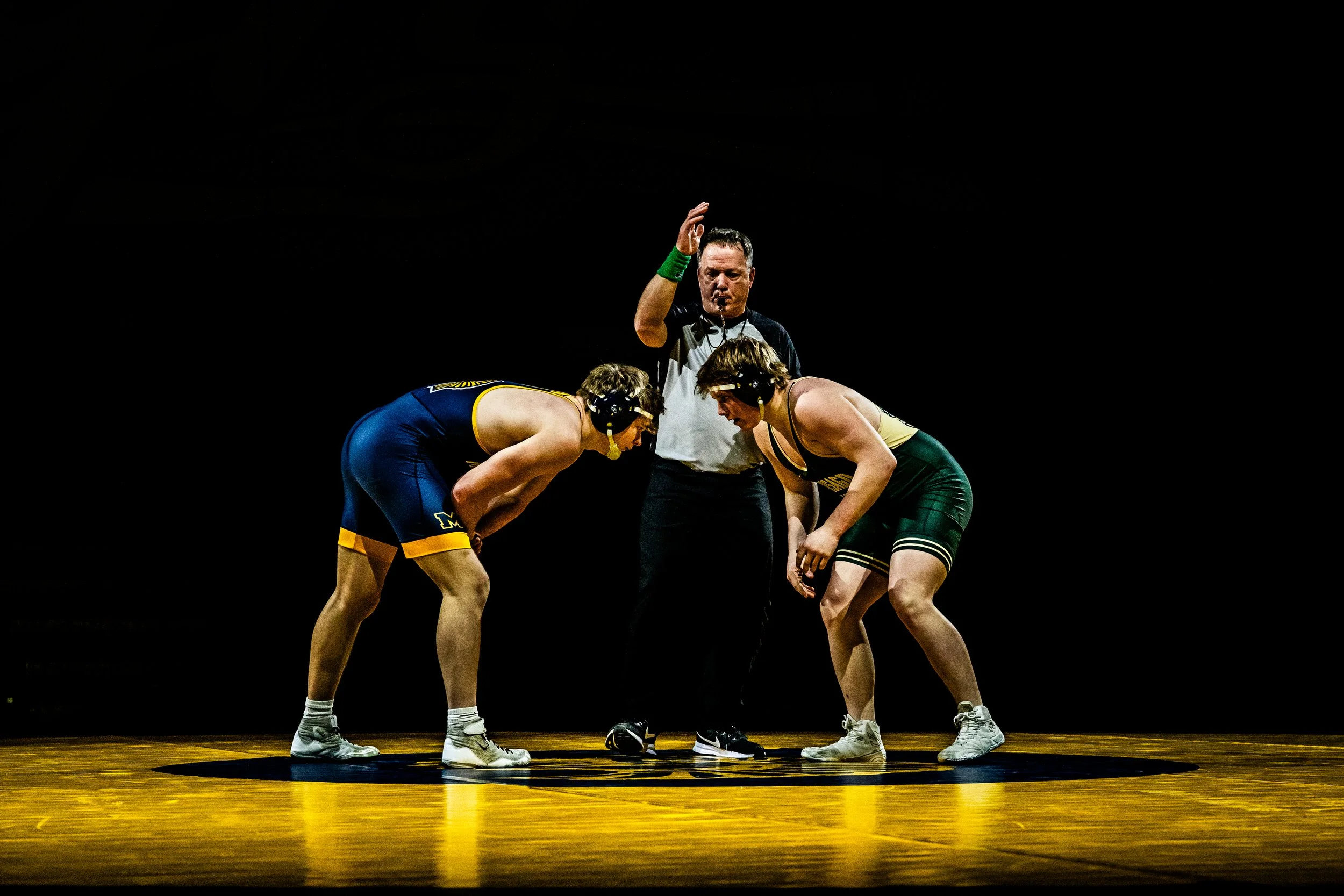 Two wrestlers in singlets and headgear face each other in a wrestling match, with a referee standing behind them, on a wooden wrestling mat with a dark background.