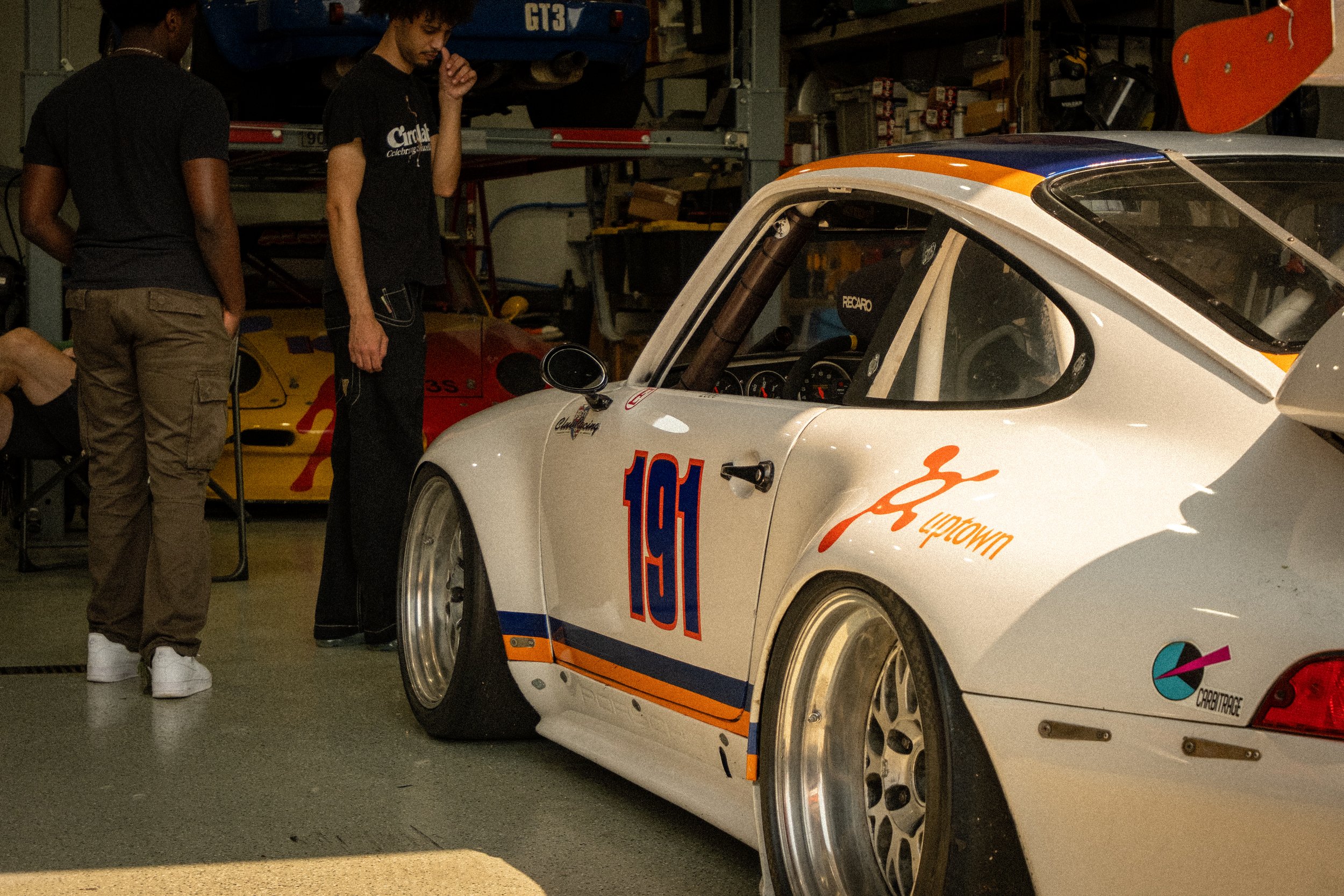 Two men are inspecting a race car with the hood open in a garage or workshop. The race car is white with orange and blue accents and has the number 105 on its side. The background contains various tools and equipment, with a yellow and red race car p