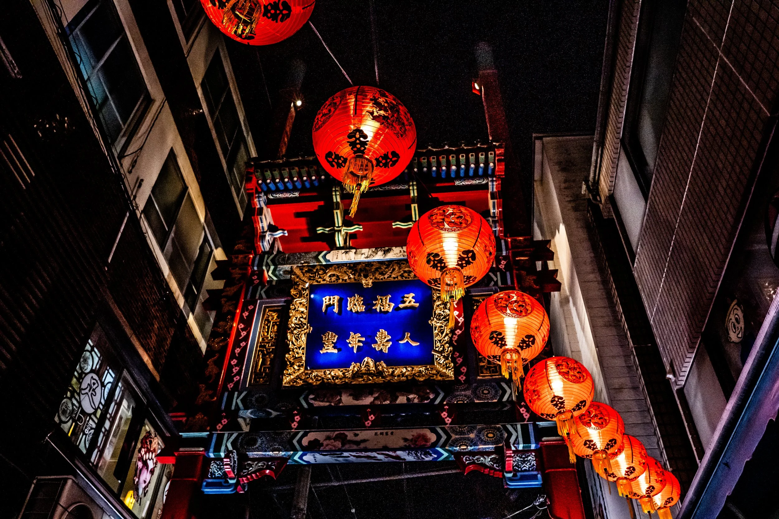 Night scene of Chinese-style lanterns hanging above a decorated entrance with Chinese characters, surrounded by building facades.